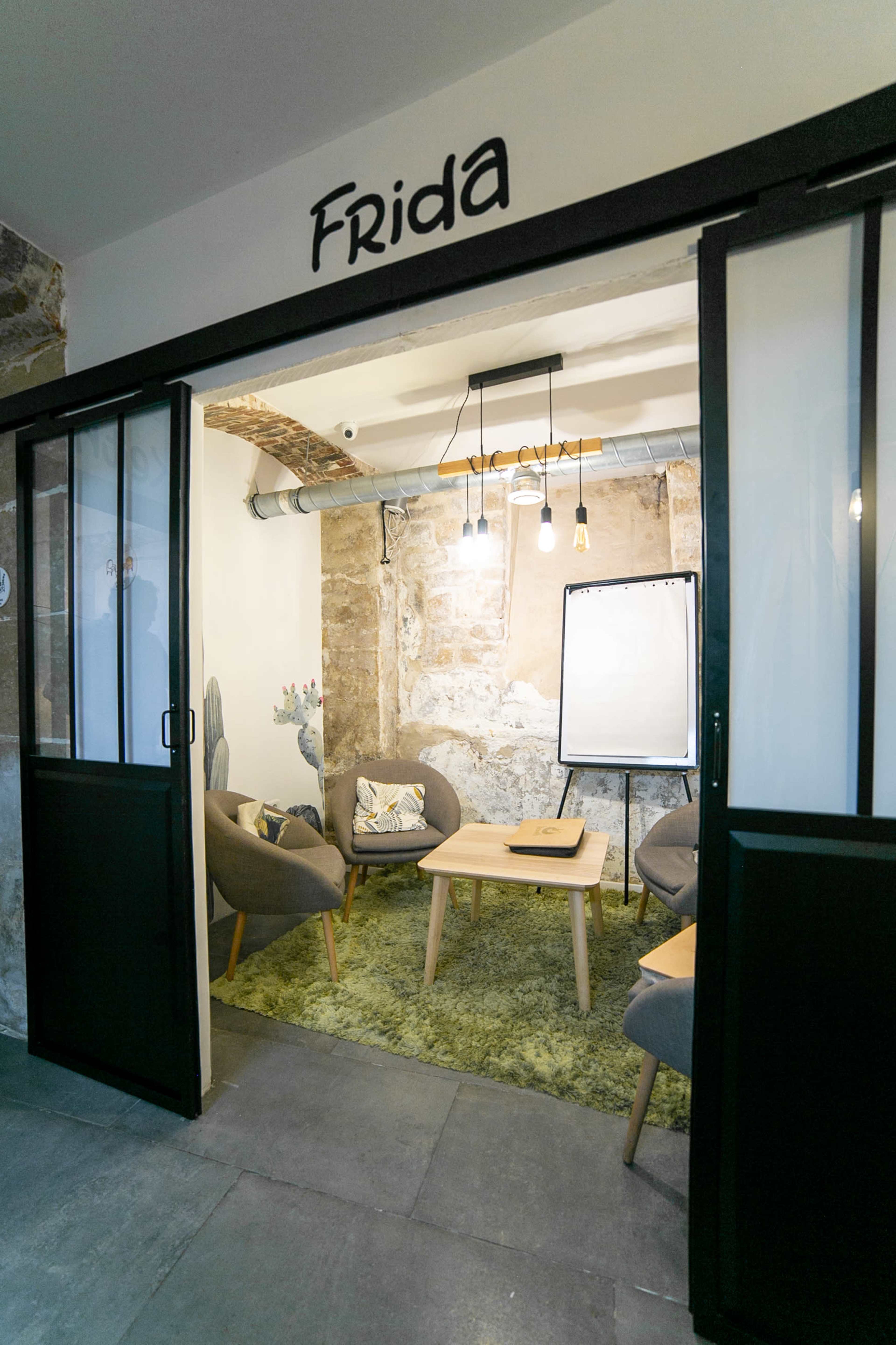 A cozy meeting space named "Frida" features a small table surrounded by gray chairs, with a whiteboard and exposed stone walls.