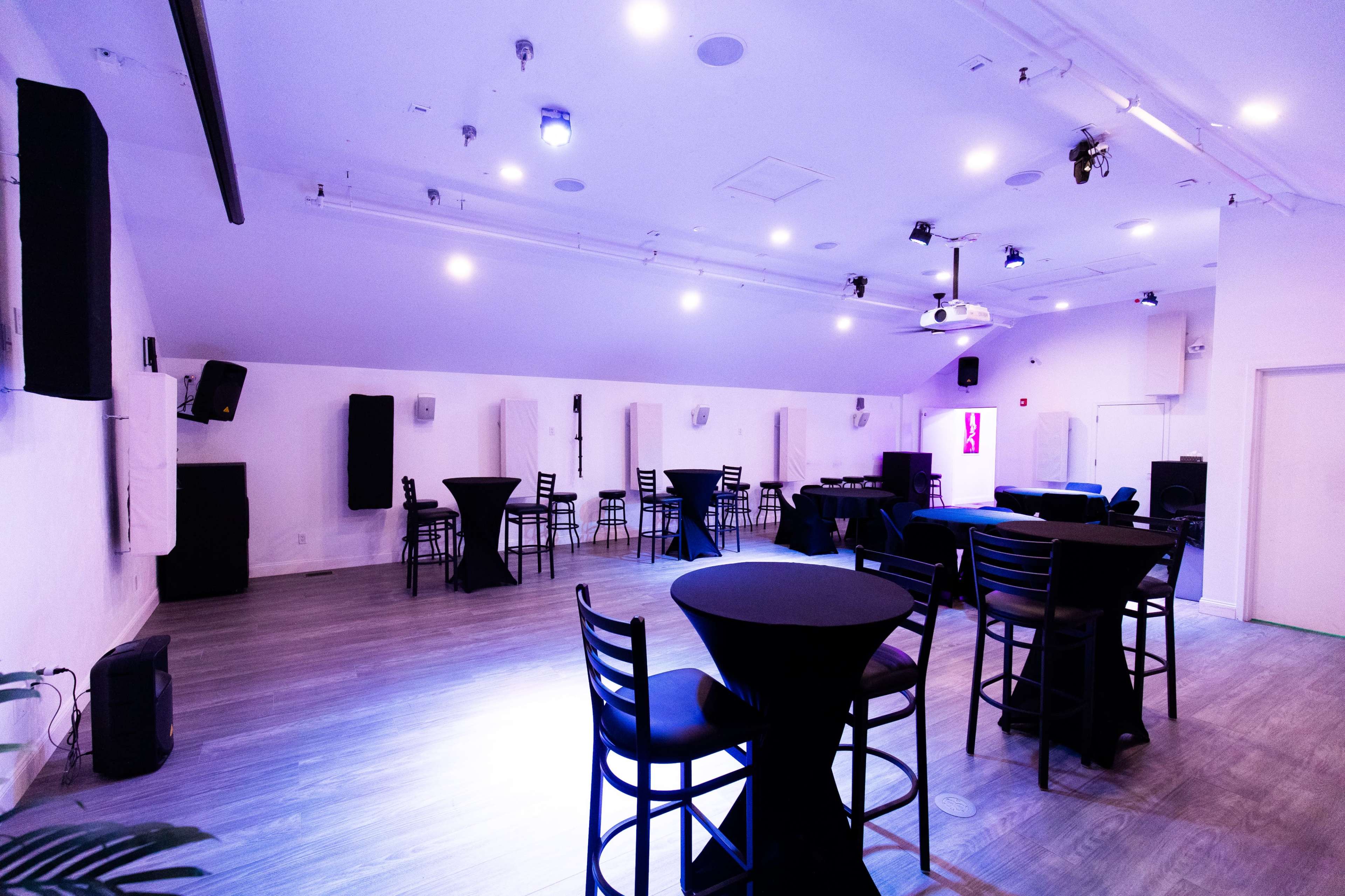 The image shows a spacious event room with high ceilings, featuring several black tables and chairs arranged around the area, along with sound equipment and wall-mounted speakers.