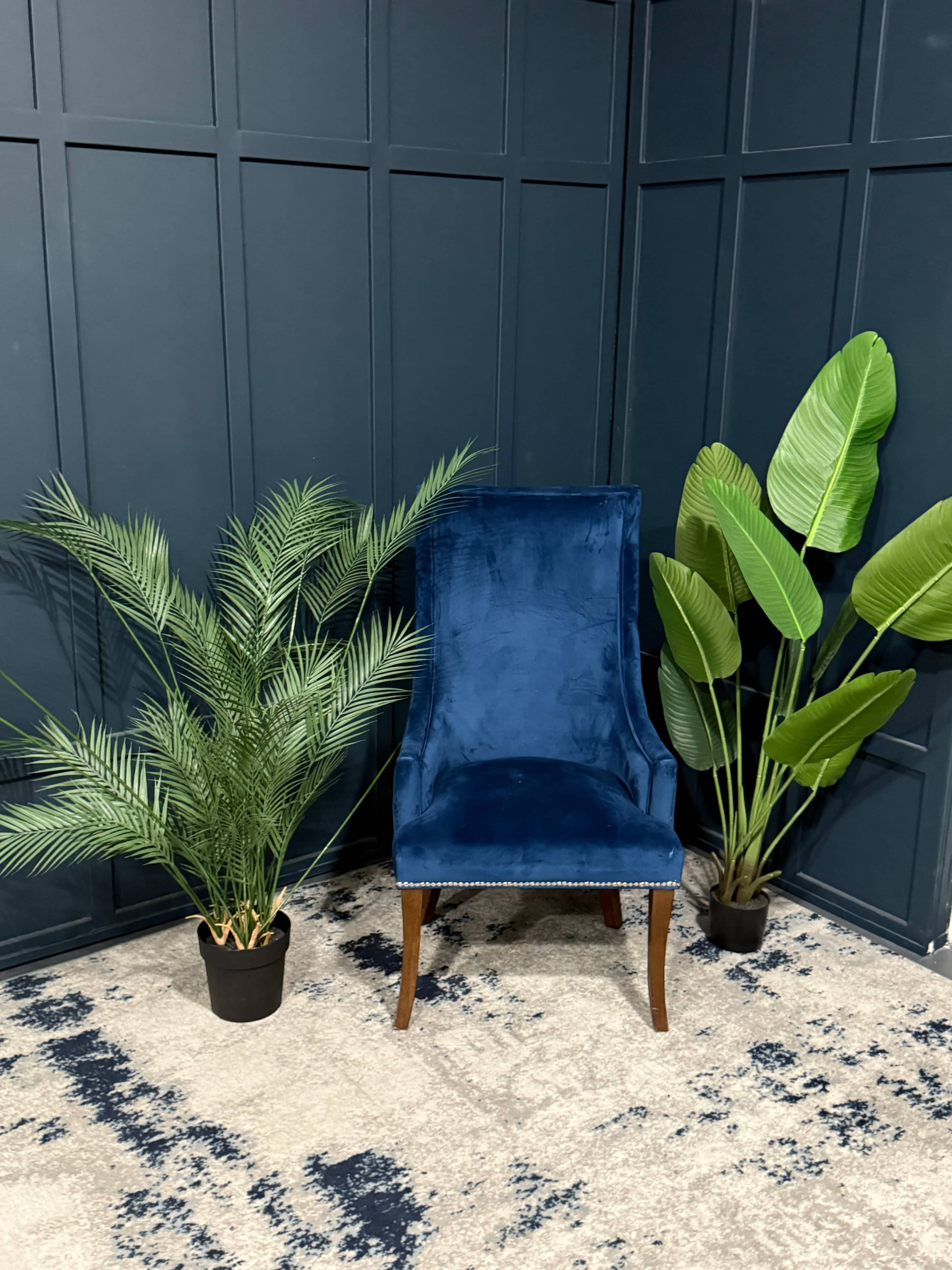 The image features a blue velvet chair positioned between two large potted plants in a room with dark green paneling and a patterned rug.