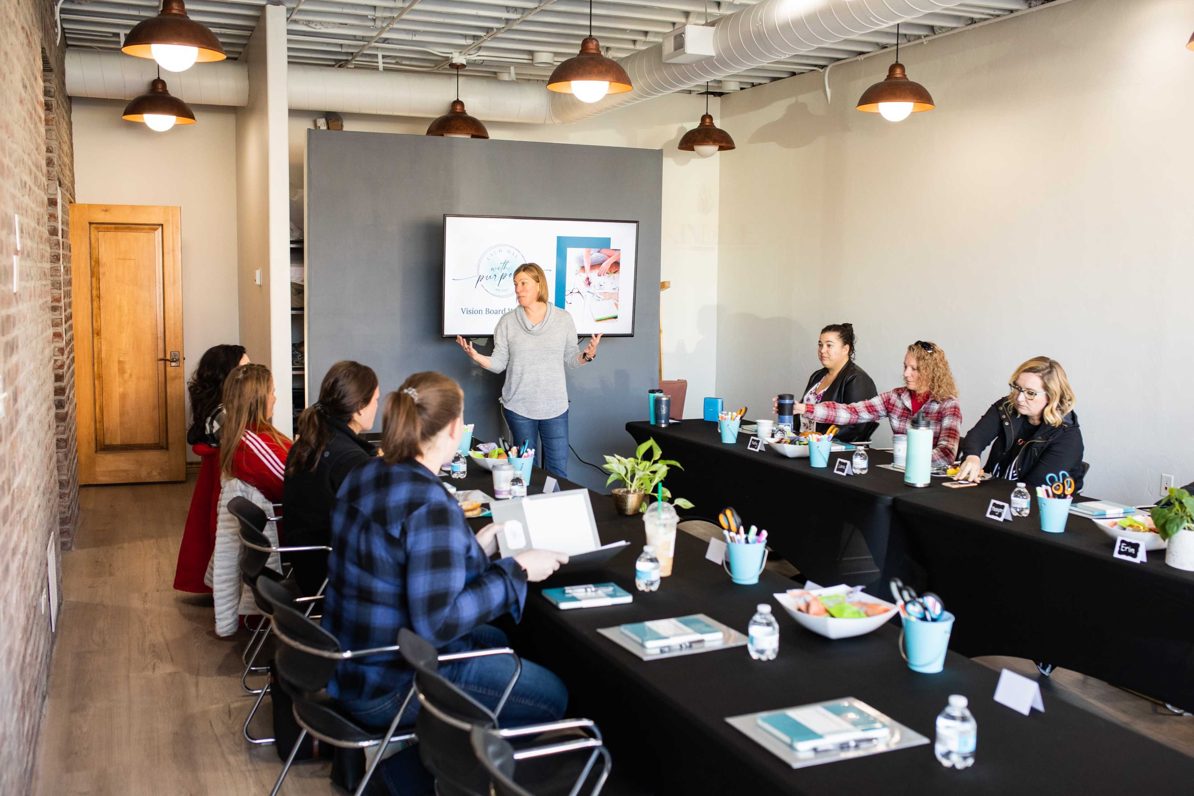 A group of people is seated at a long table in a meeting room, while a woman stands at the front presenting on a screen.