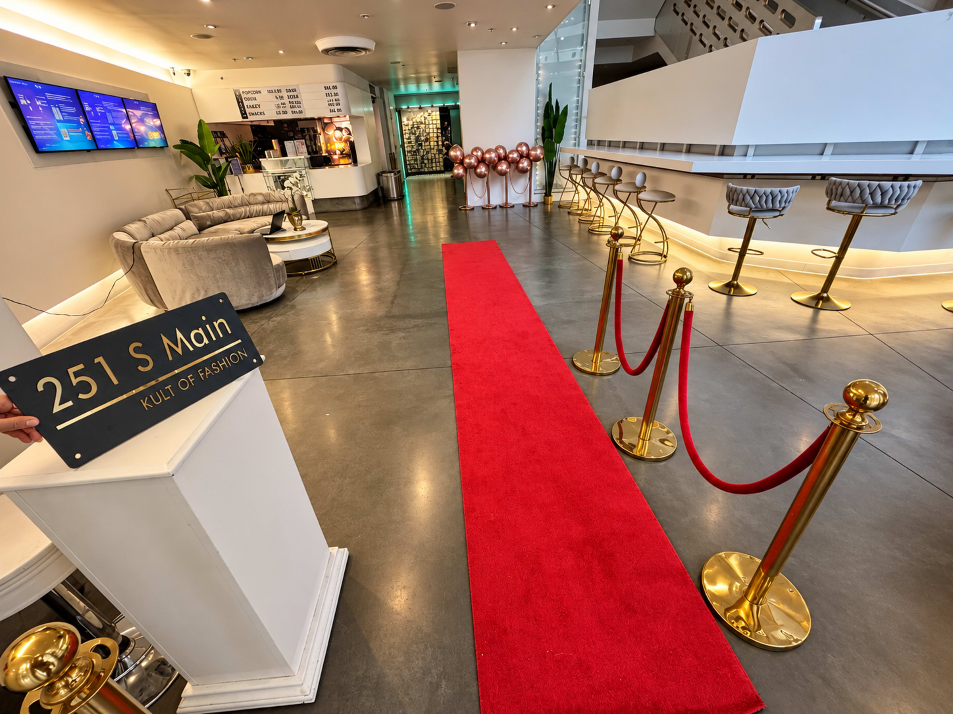 The image shows a modern lobby area featuring a red carpet leading to a seating area, with a welcome sign displaying "251 S Main" and a row of bar stools.