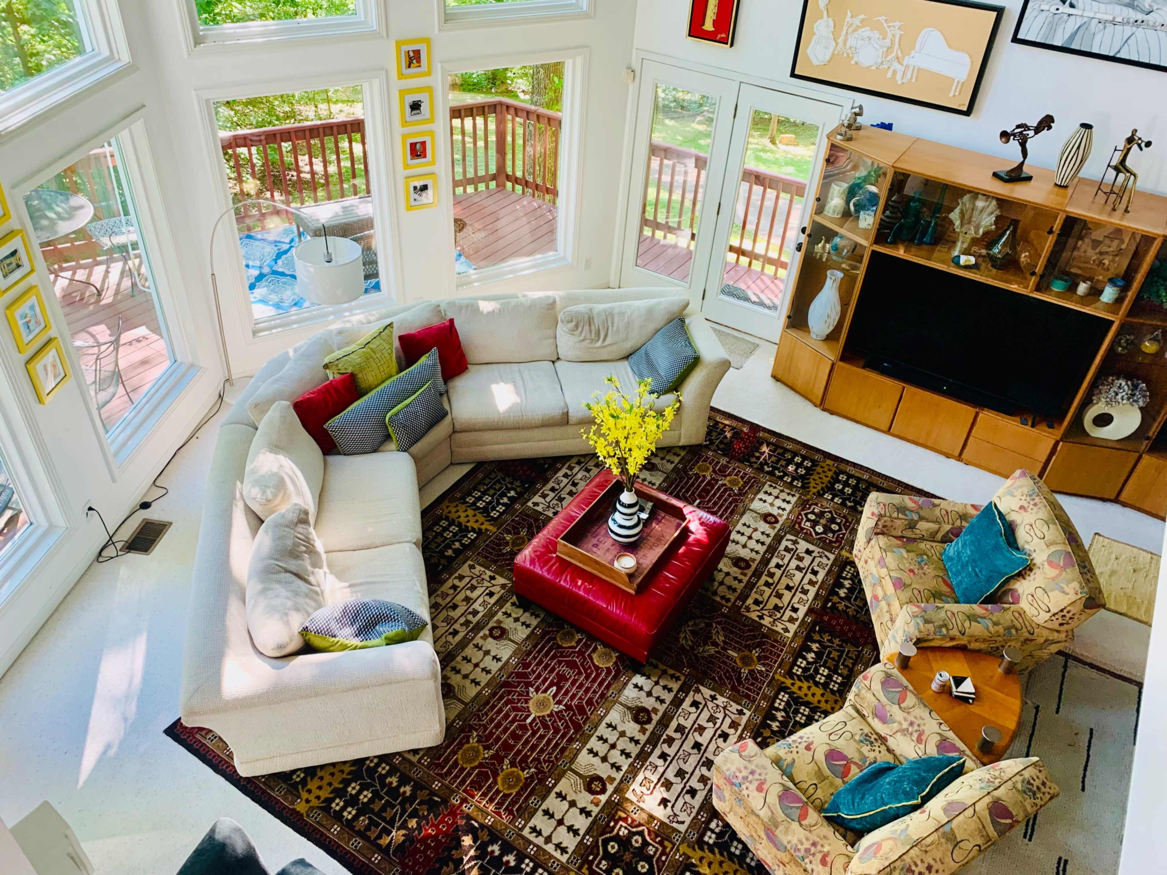 The image shows a bright living room with a sectional sofa, two patterned chairs, a coffee table, and large windows overlooking a deck.