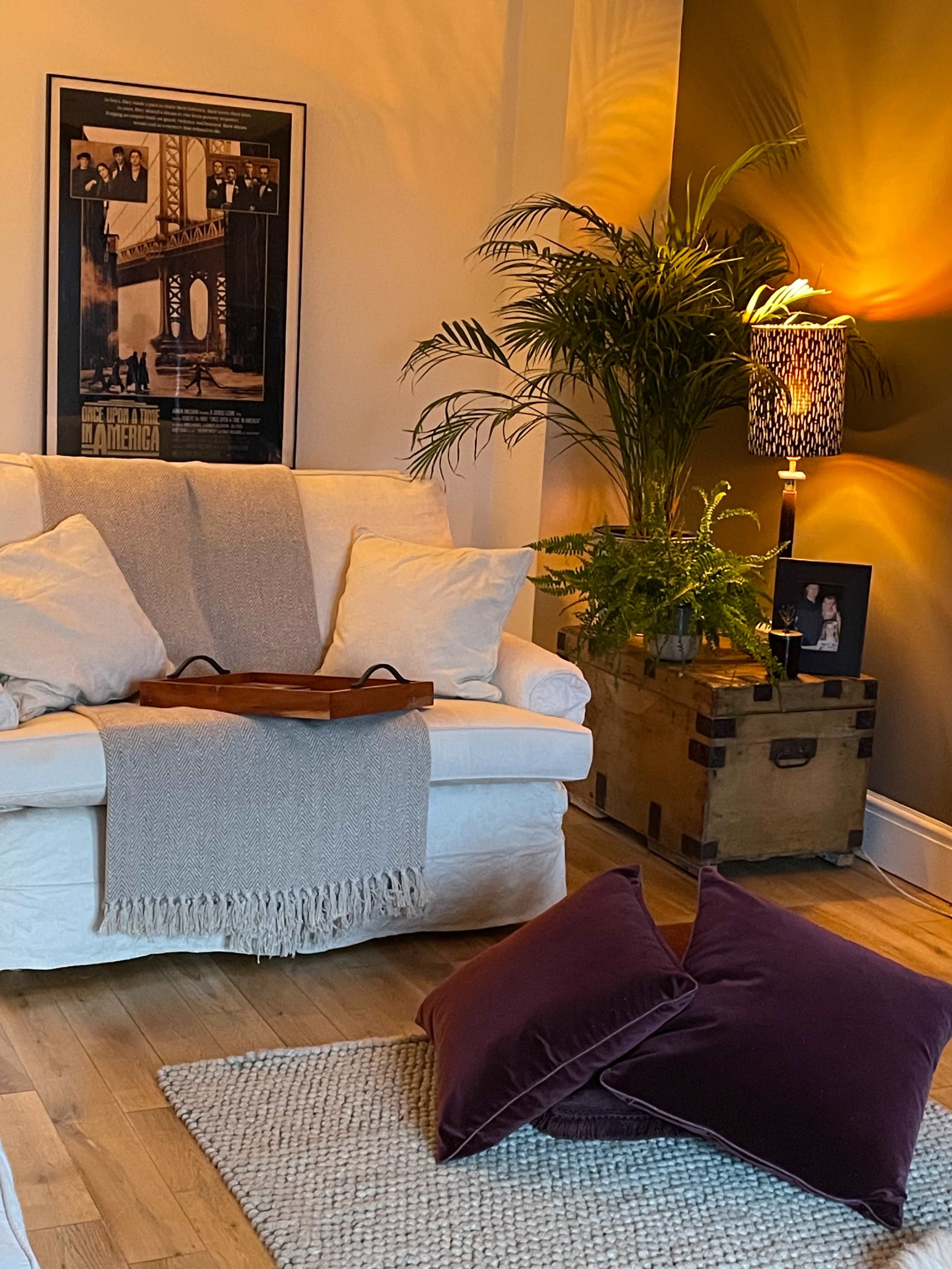 A cozy living room features a white sofa with decorative pillows, a wooden tray on its arm, a green plant beside a lamp, and a vintage poster on the wall.
