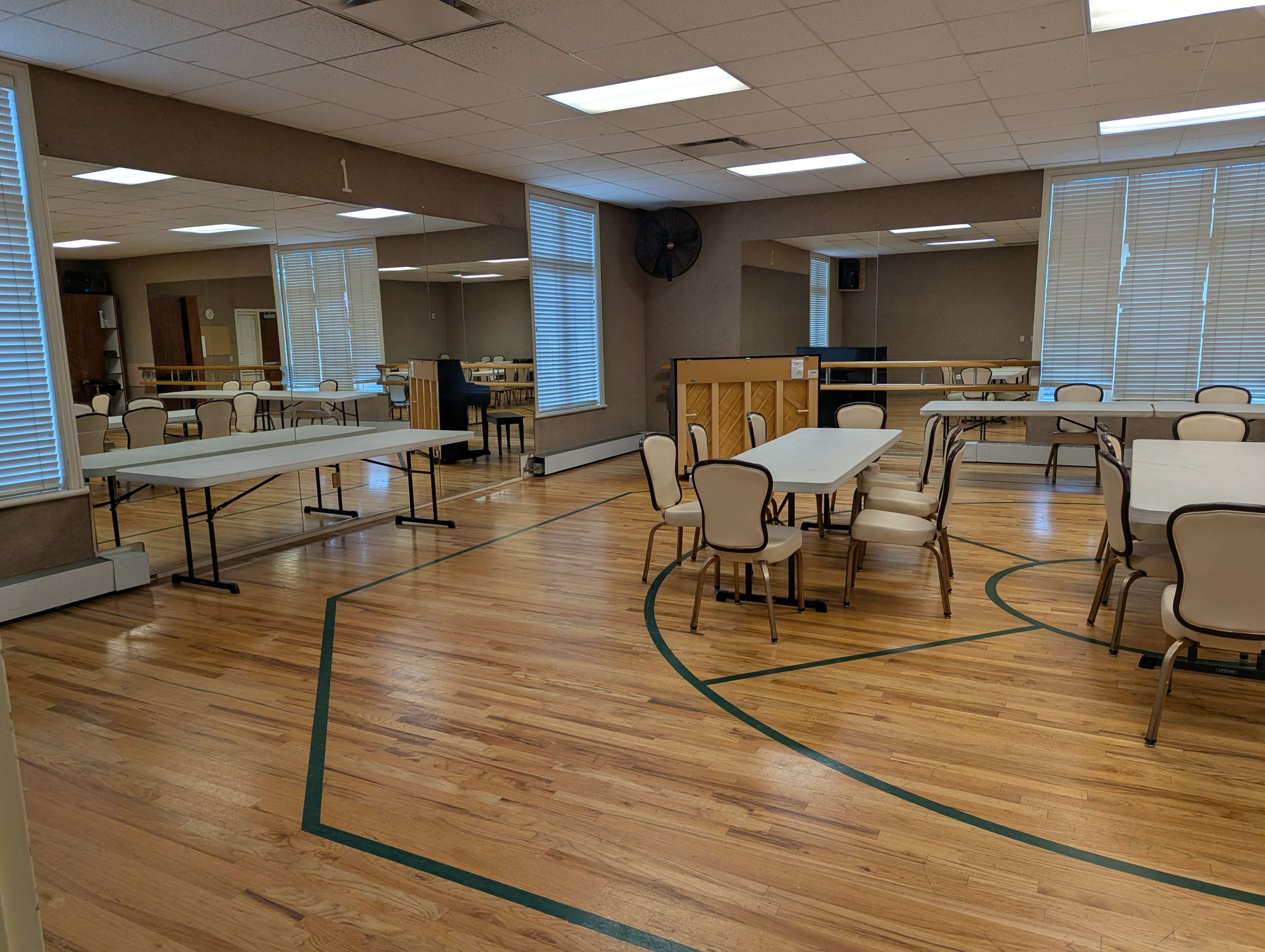 An empty room features several tables and chairs arranged around a polished wooden floor with mirrored walls and windows providing natural light.