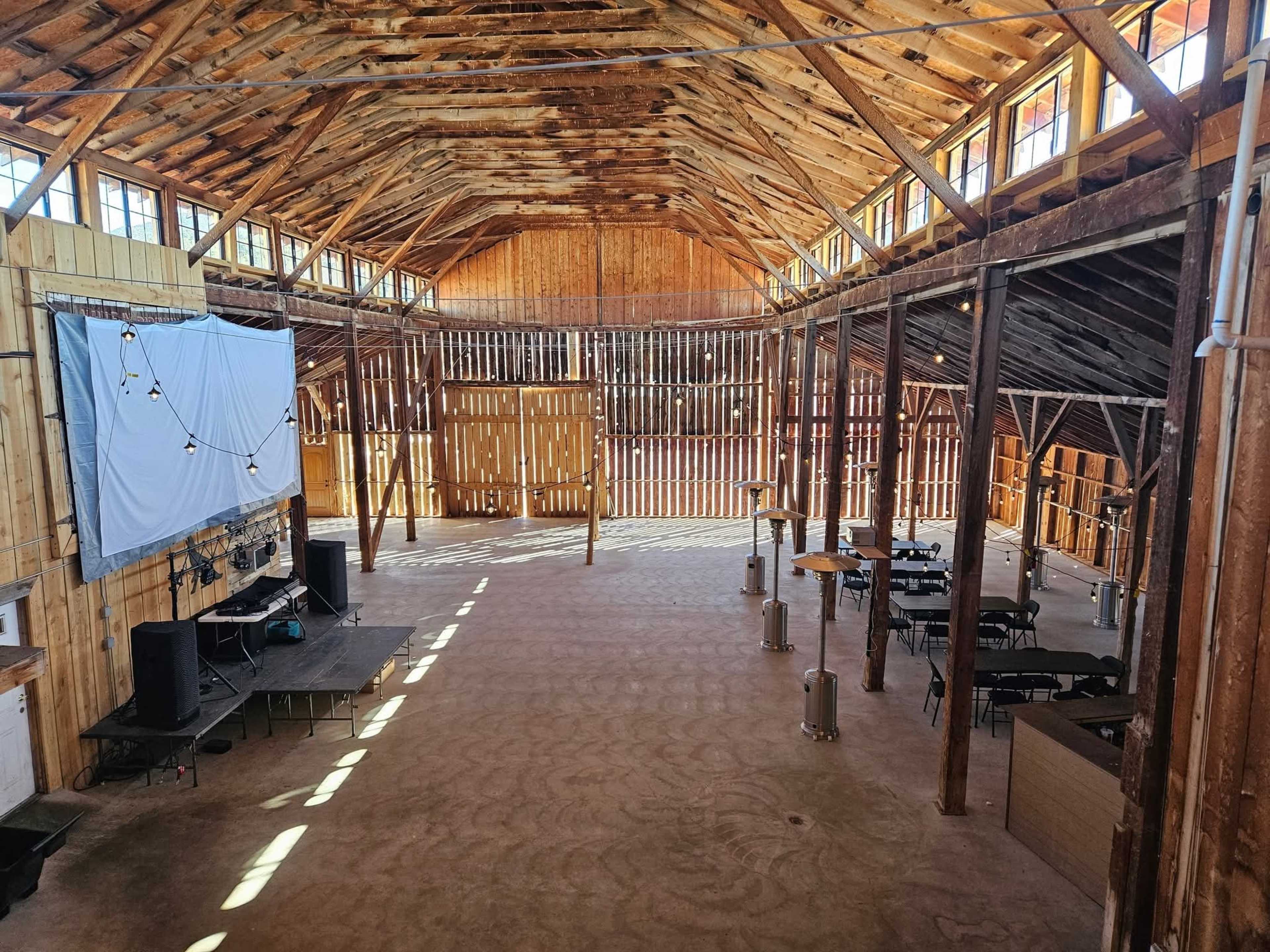 The image shows a spacious wooden barn interior with high ceilings, wooden beams, and a stage setup at one end.