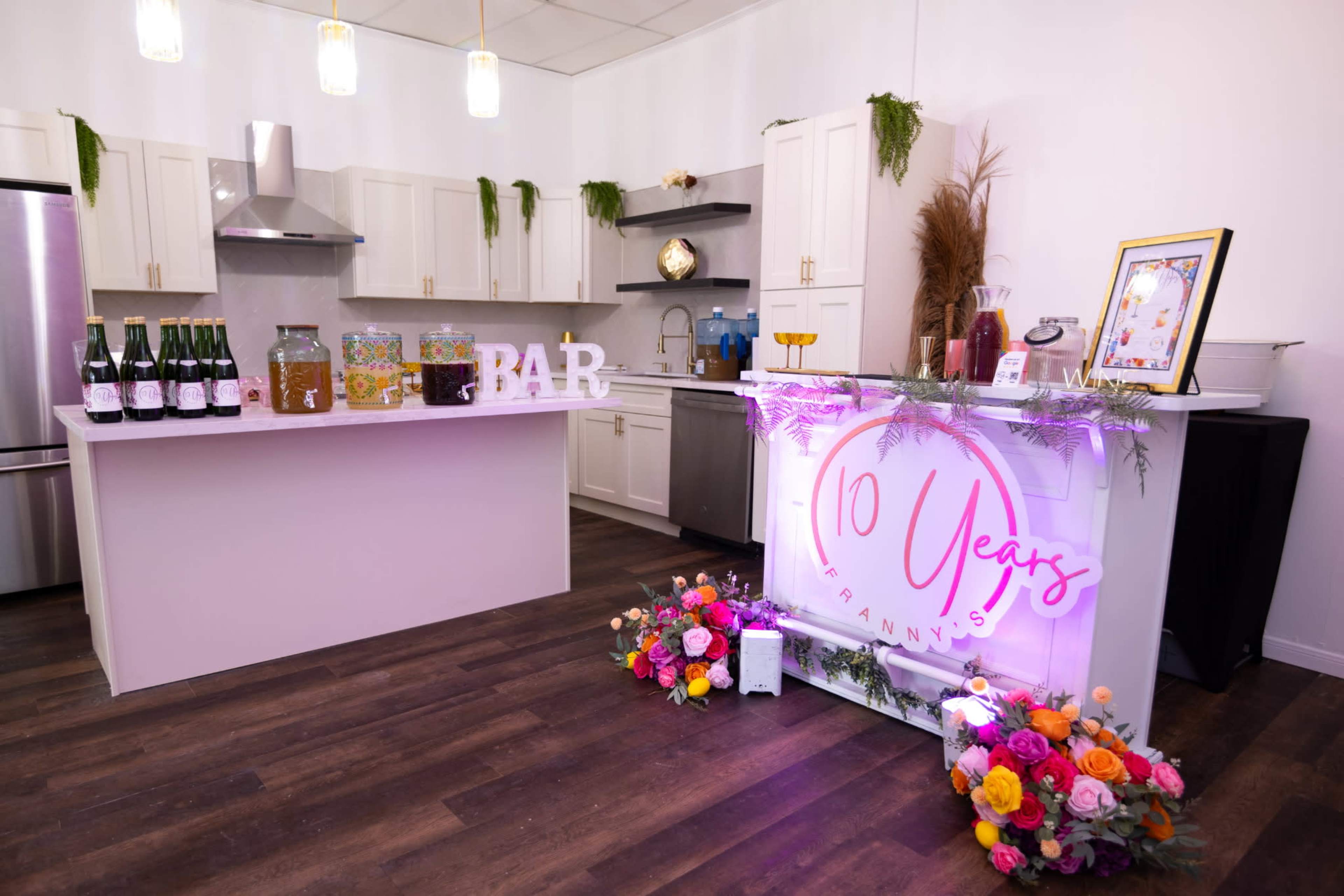 The image shows a decorated kitchen space featuring a bar with colorful drinks, a celebration sign reading "10 Years," and floral arrangements.