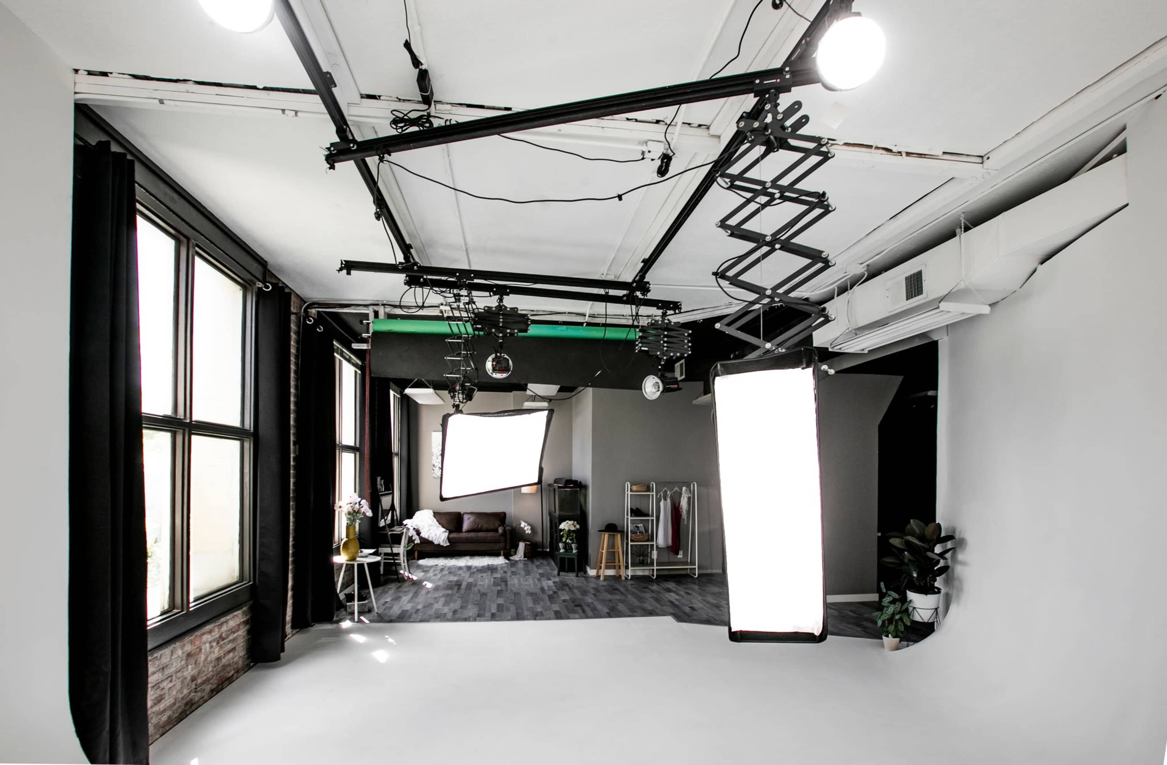 A spacious photography studio with large windows, lighting equipment suspended from the ceiling, and a plain backdrop.
