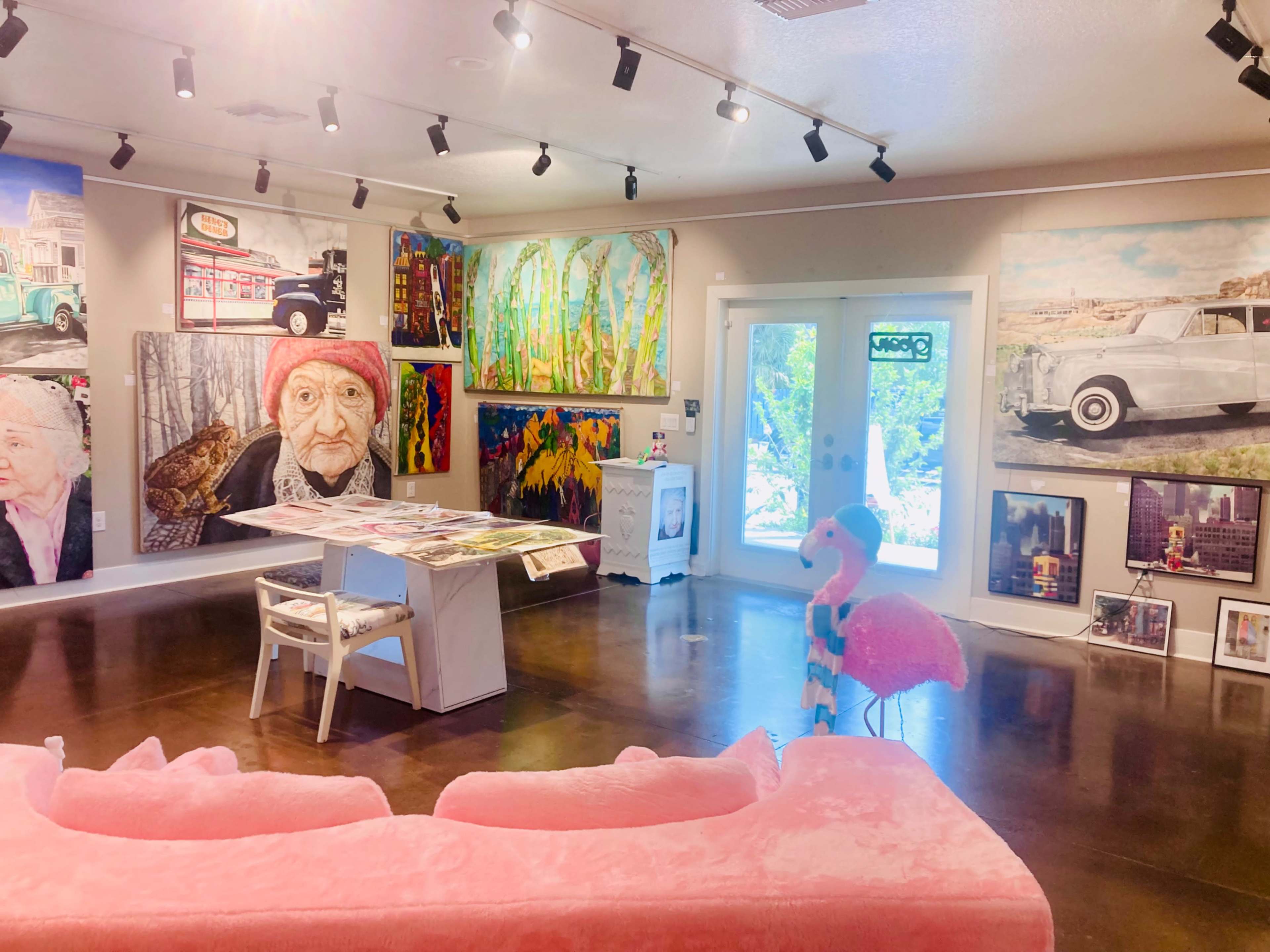 The image shows an art gallery interior featuring various large paintings on the walls, a pink sofa, and a table in the center with artworks displayed on it.