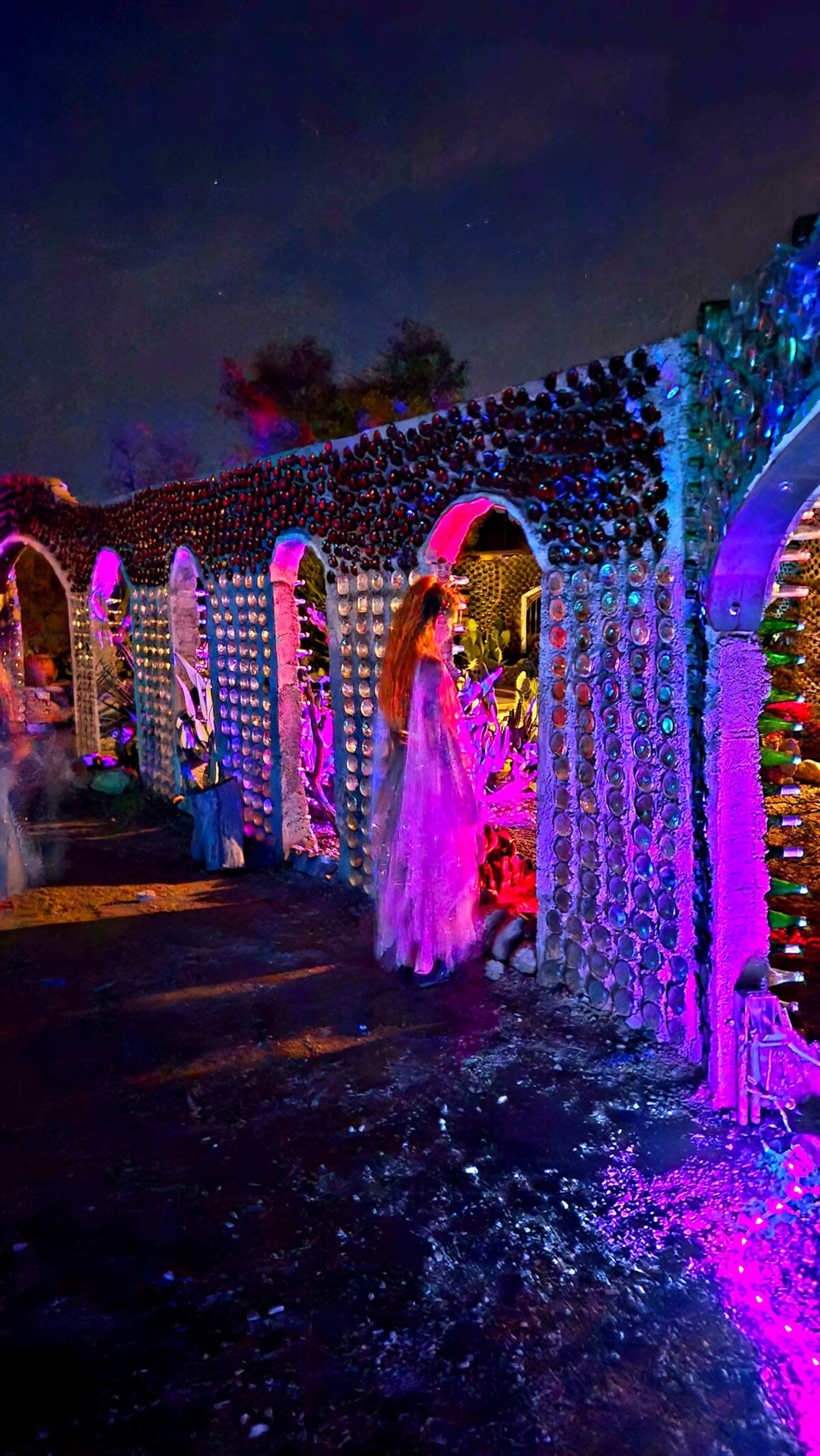 The scene depicts an illuminated outdoor walkway with arches made of bottles, surrounded by colorful lights and decorations.