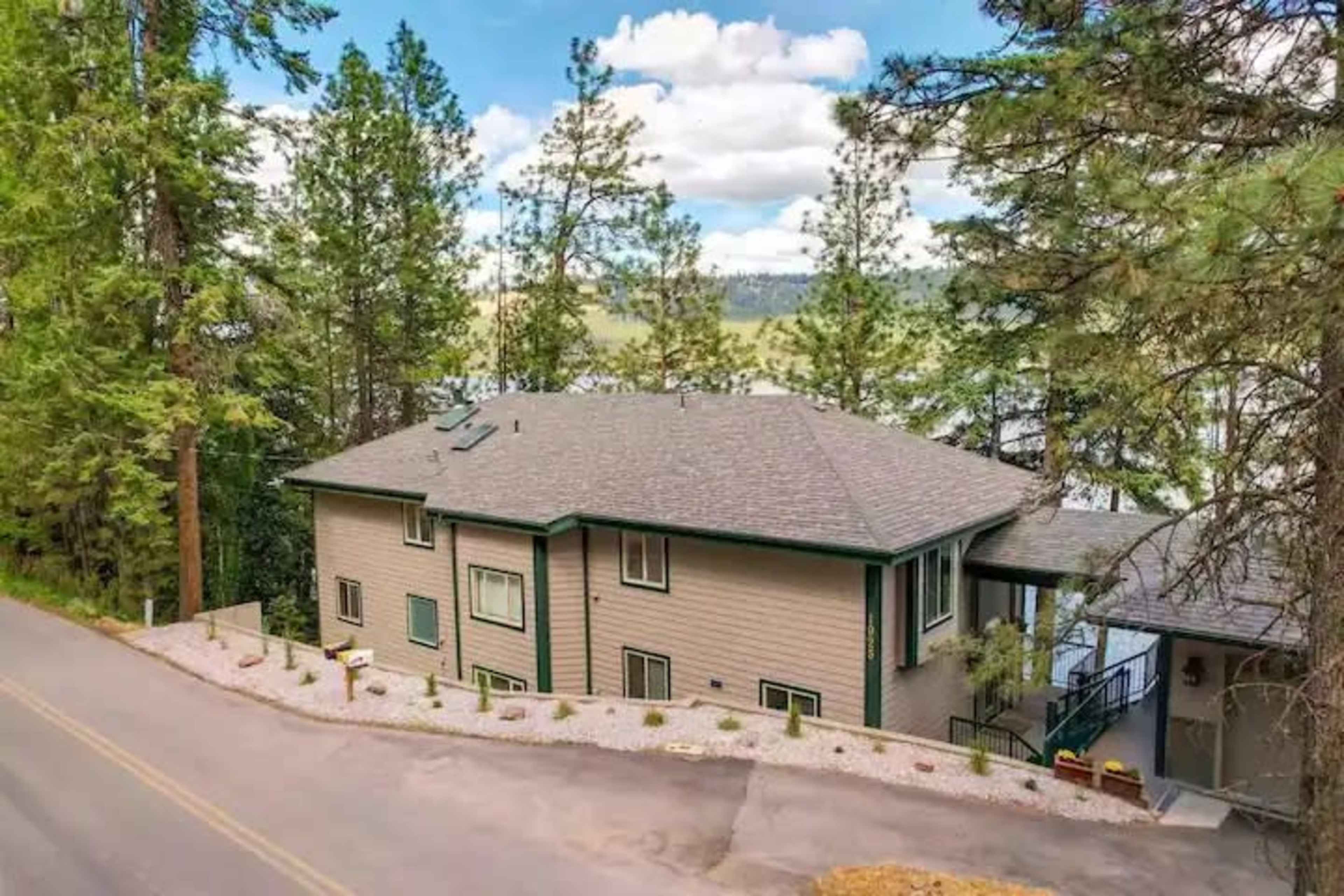 A large two-story house is located on a hillside next to a lake, surrounded by tall pine trees.