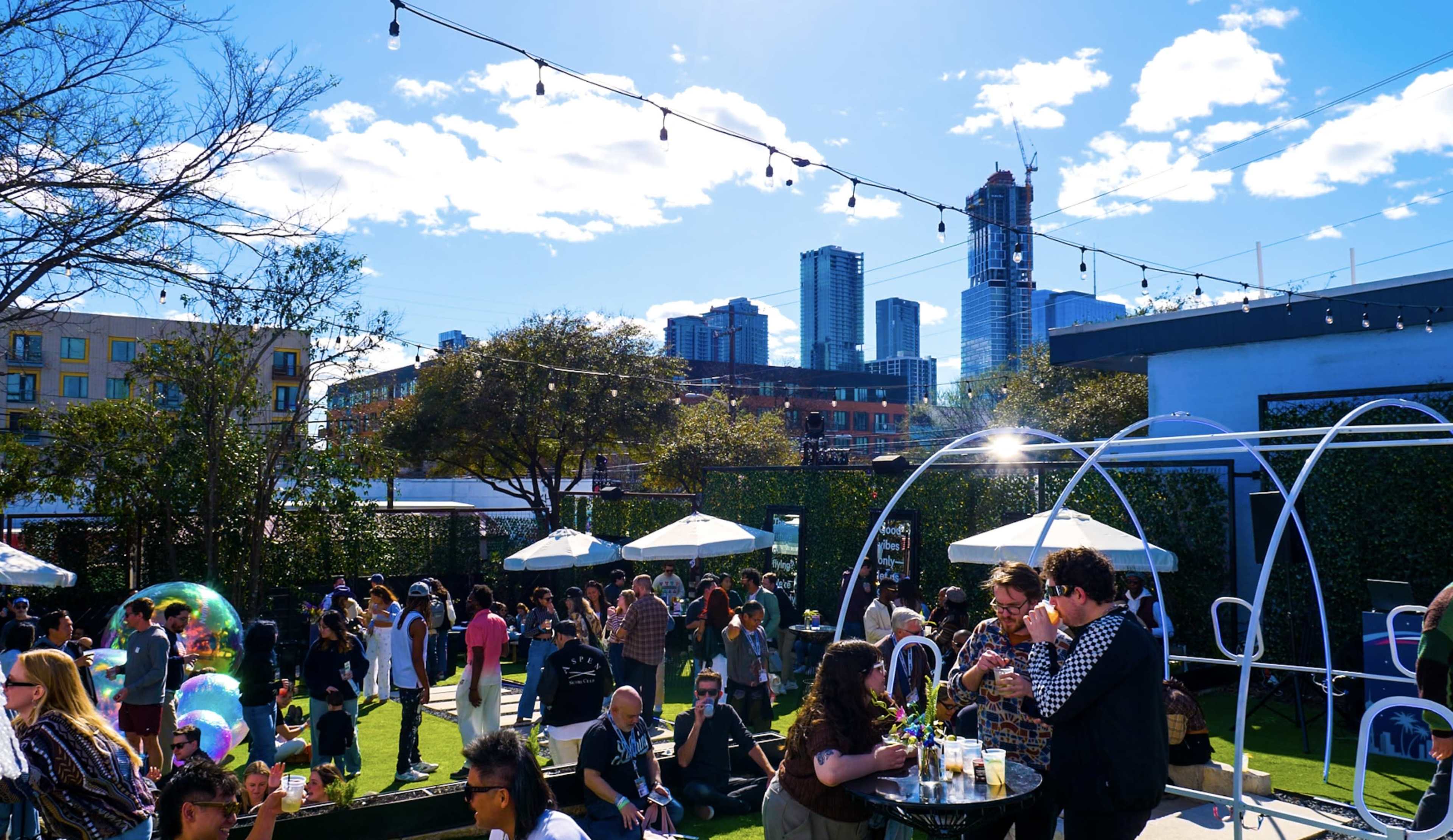 A lively outdoor gathering takes place on a rooftop, featuring people socializing, string lights overhead, and a skyline of tall buildings in the background.