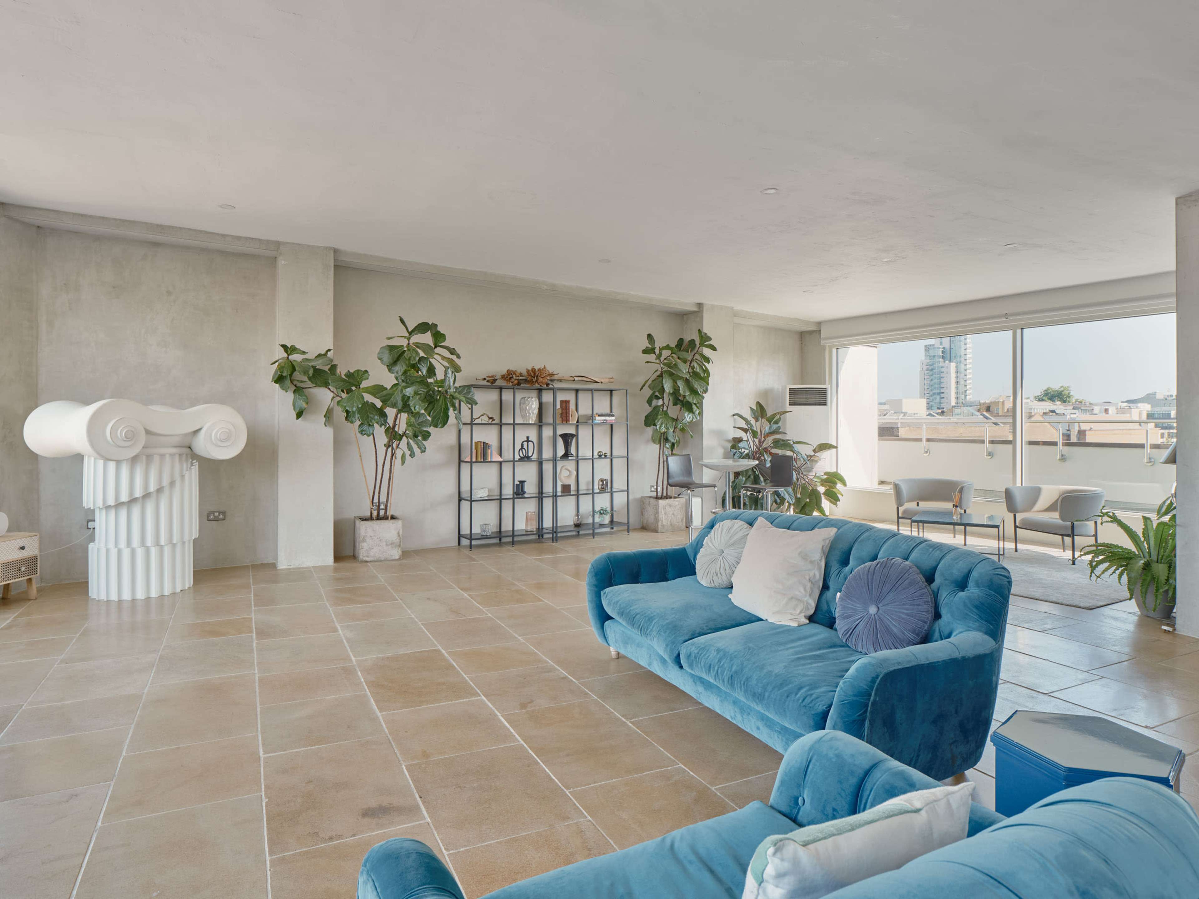 The image shows a spacious modern living area with blue sofas, a bookshelf, potted plants, and large windows providing natural light.