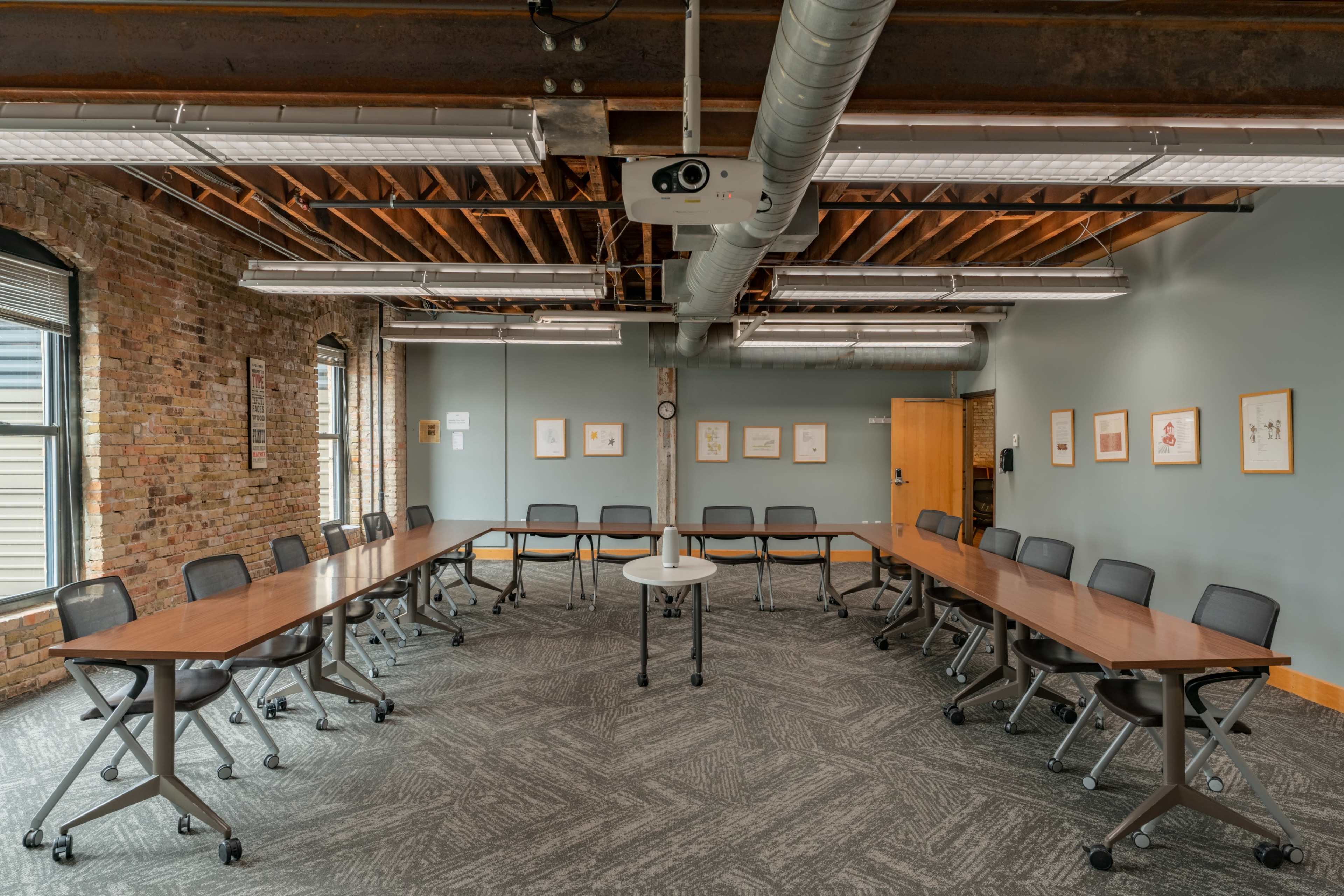 A spacious meeting room features a U-shaped arrangement of tables and chairs with exposed brick walls and overhead lighting.