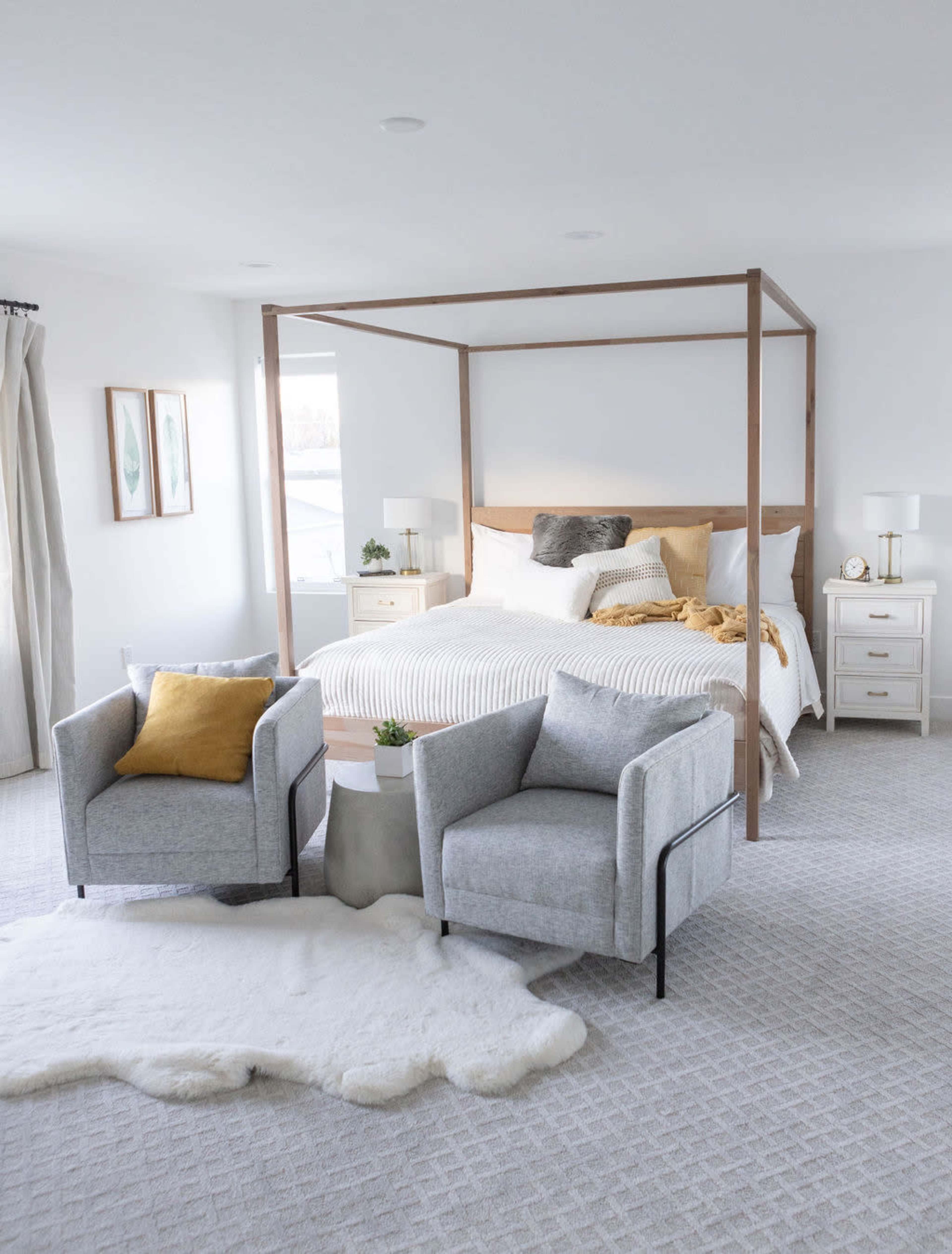 A modern bedroom features a four-poster bed with light bedding, two gray chairs, and a small table on a textured rug.
