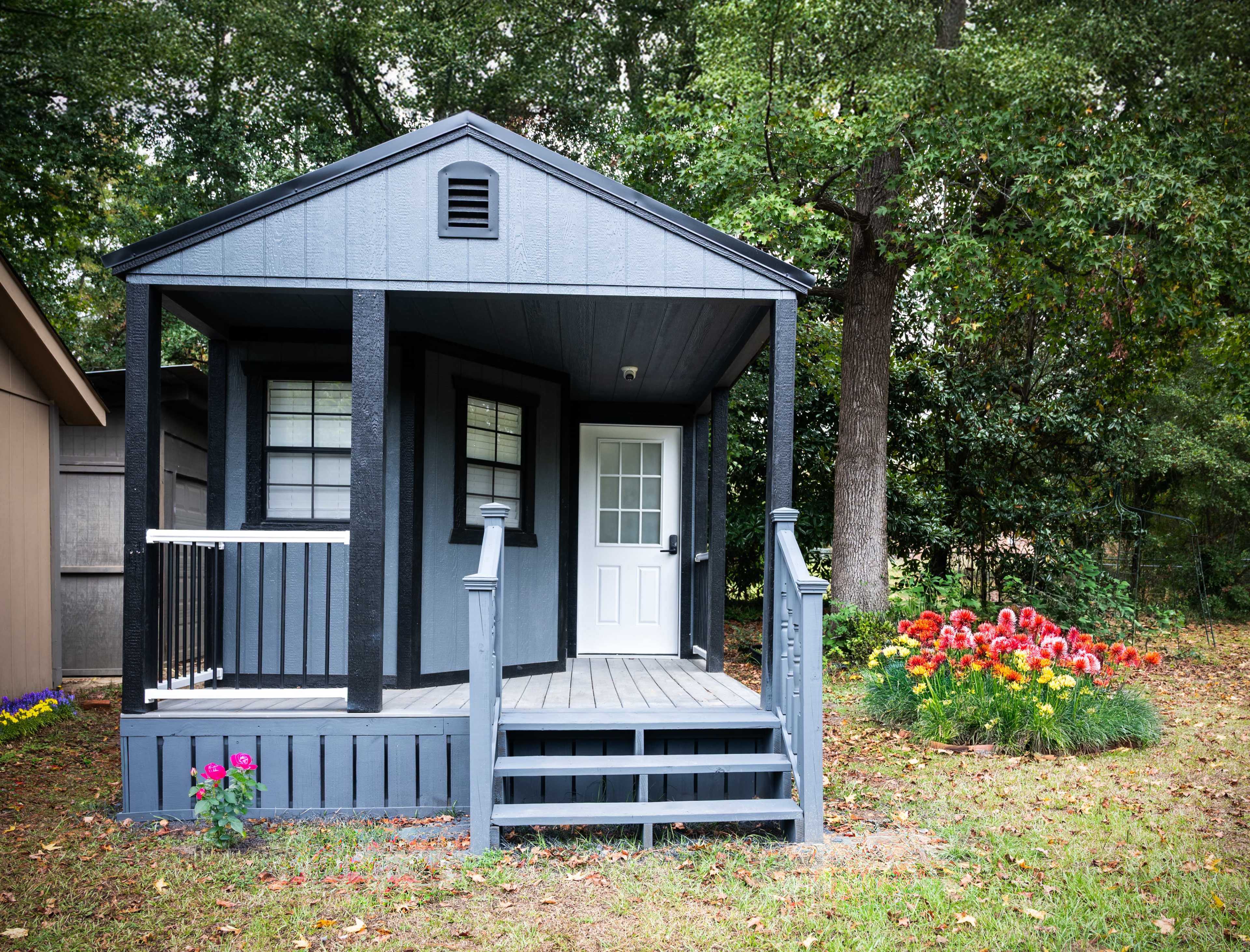 The image shows a small, gray cottage with a porch, steps leading up to the front door, and colorful flower beds nearby.