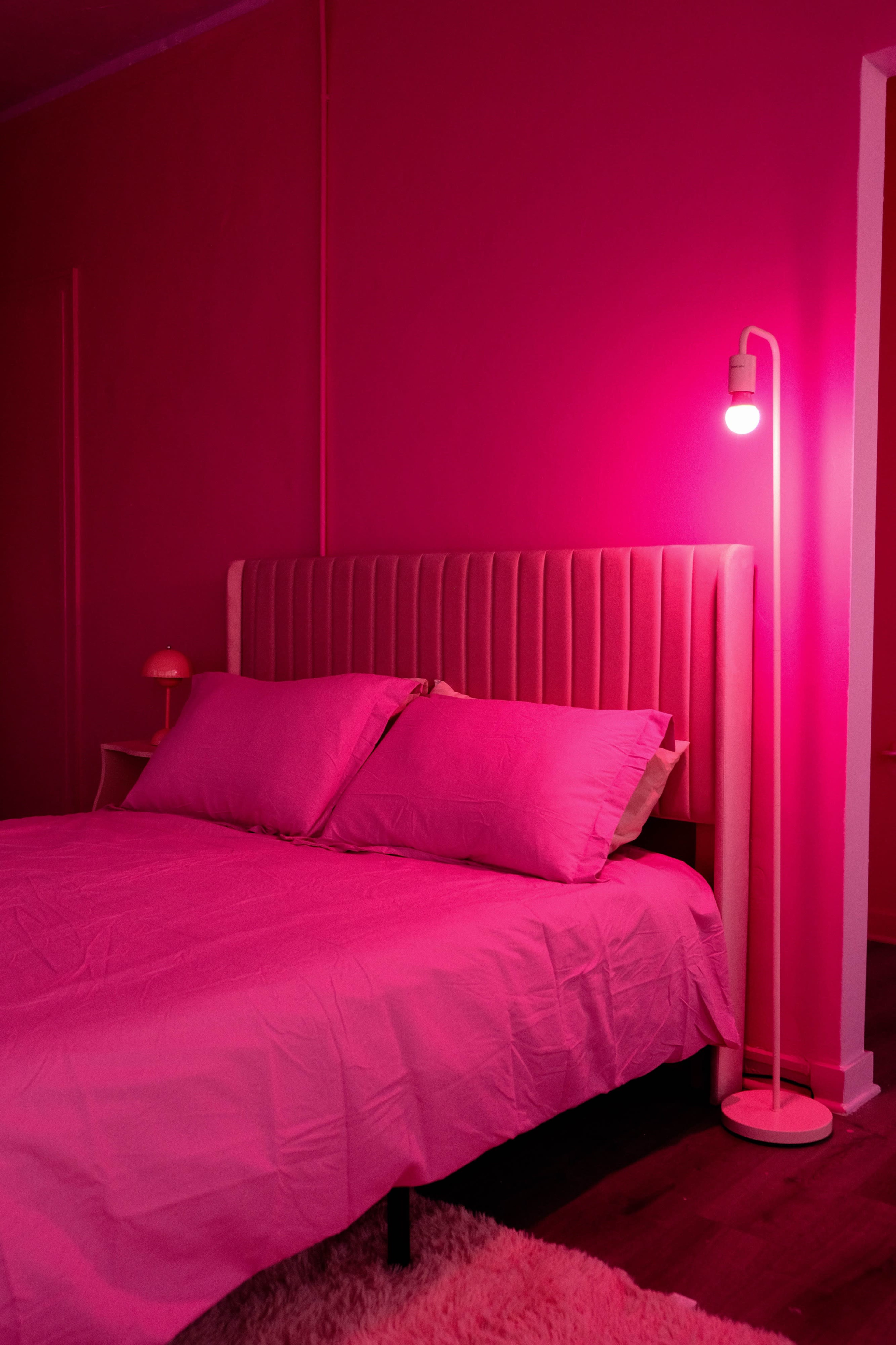 The image shows a bedroom with pink walls, a bed with pink bedding, and a pink lamp beside the bed.