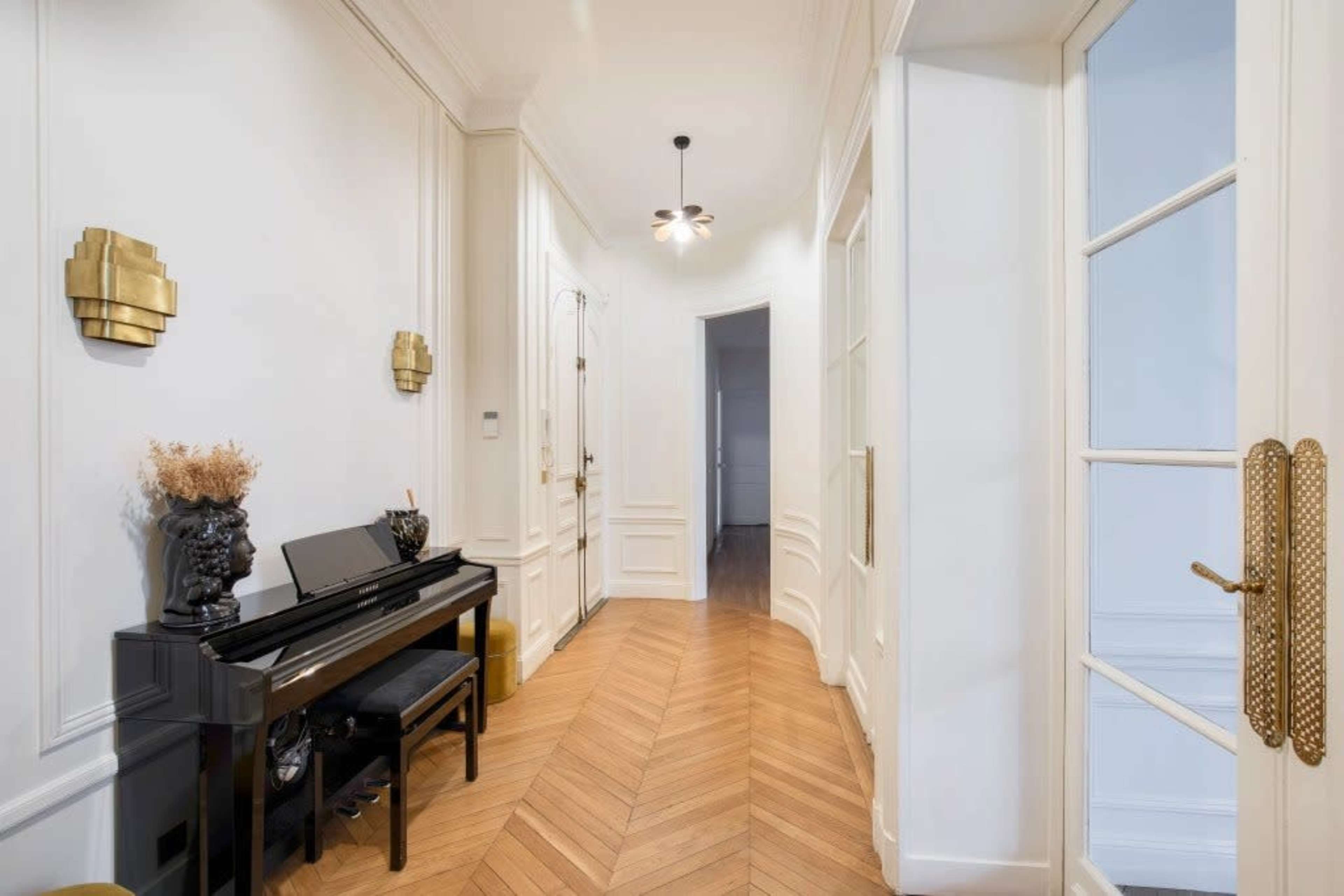 The image shows a hallway featuring a black piano against a white wall, with wooden flooring and stylish decorative elements.