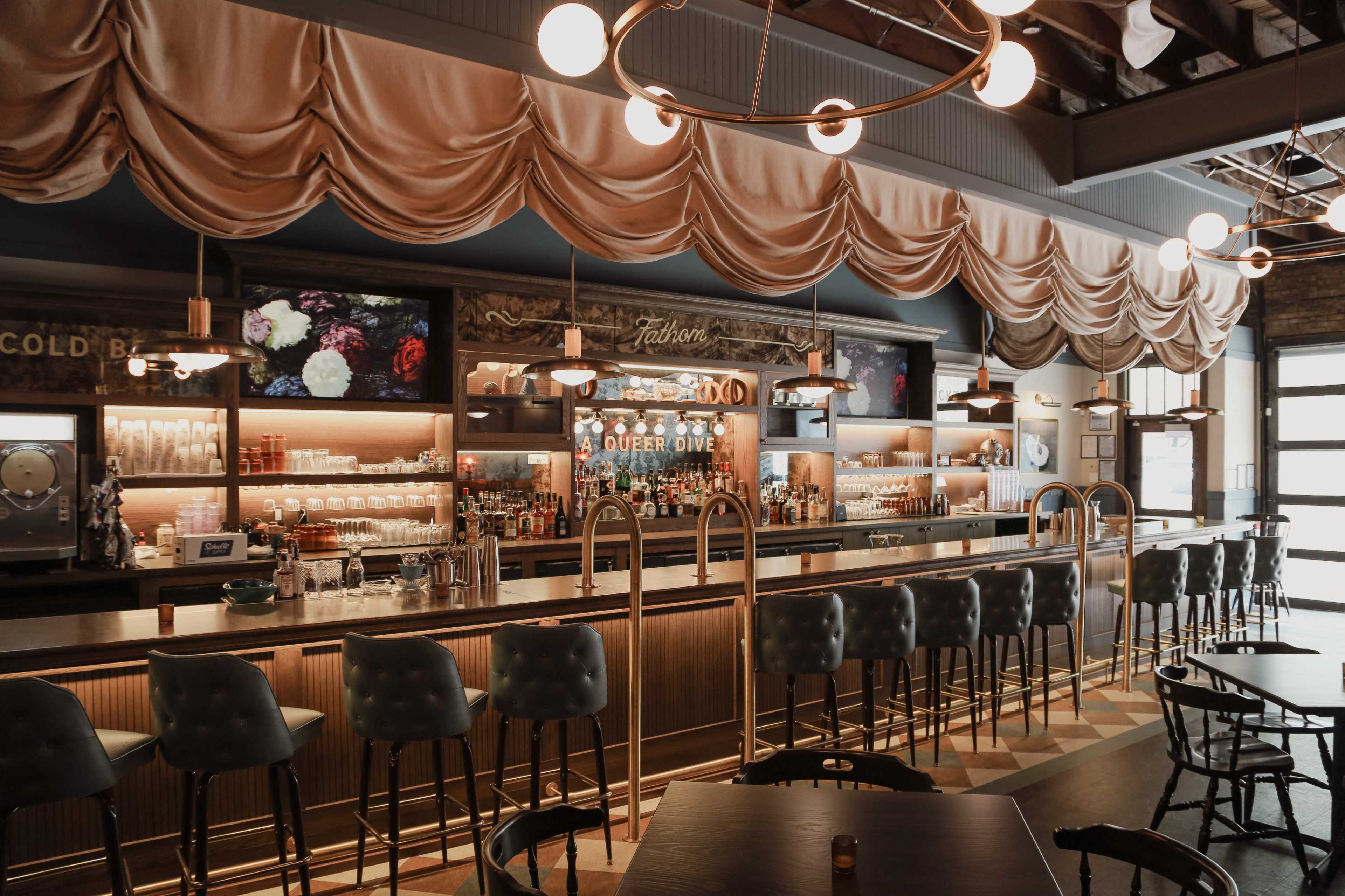 The image shows a modern bar with a long counter, elegant draped fabric above, and shelves stocked with various bottles and glassware.