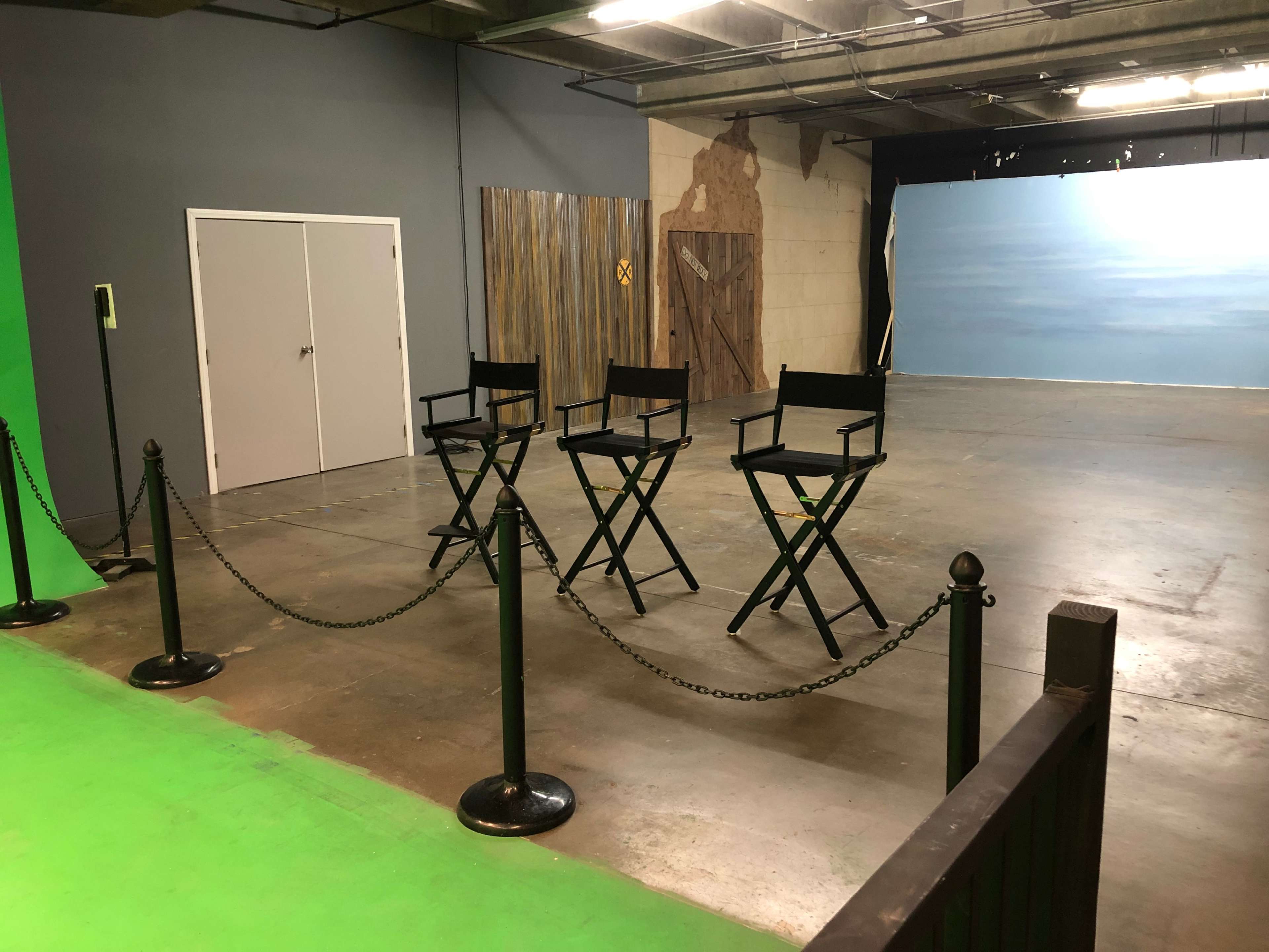 A sparse studio space with three black director's chairs arranged in front of a painted backdrop, surrounded by a chain barrier.