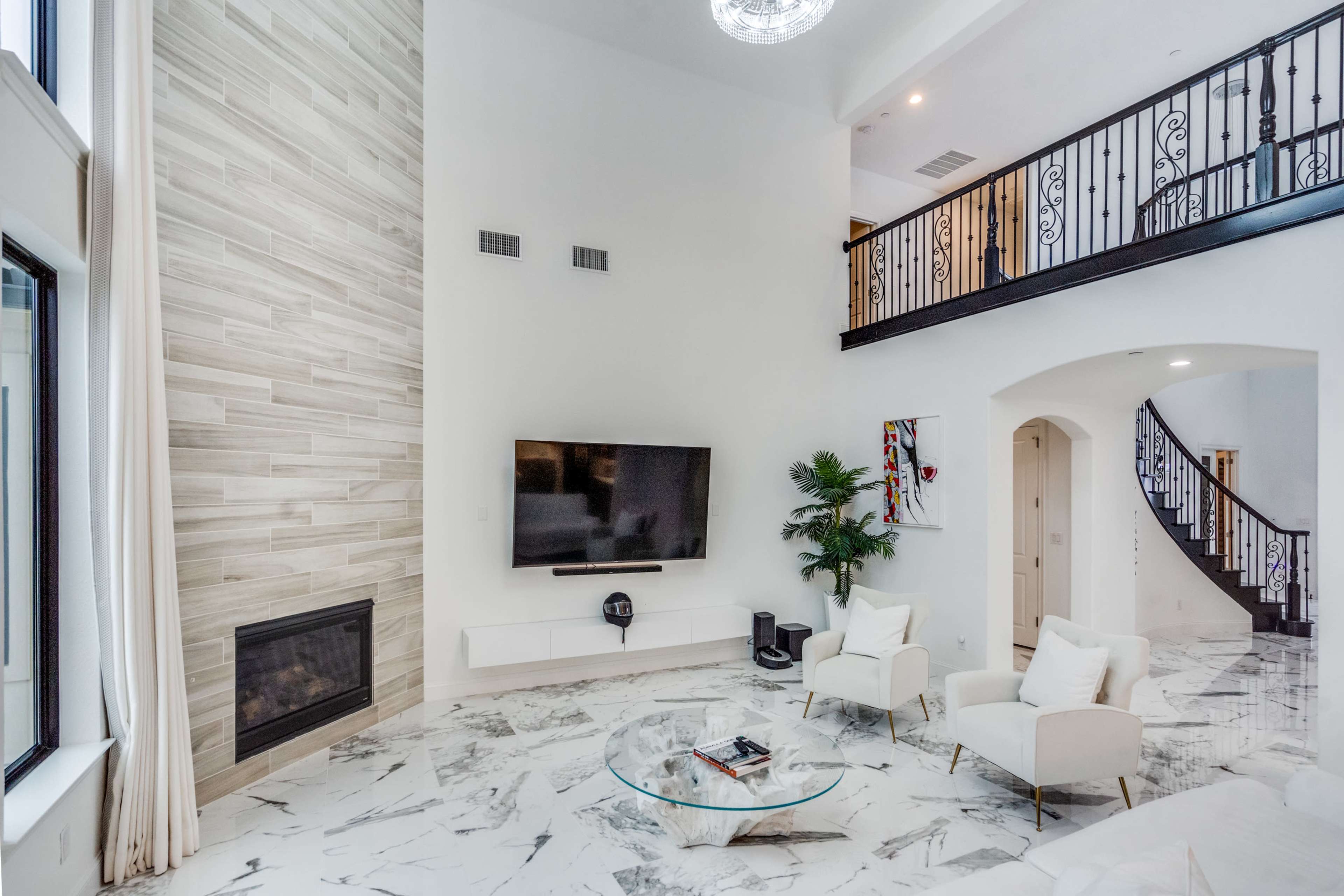 The image shows a modern living room with a high ceiling, a large television mounted on the wall, a glass coffee table, and marble flooring.