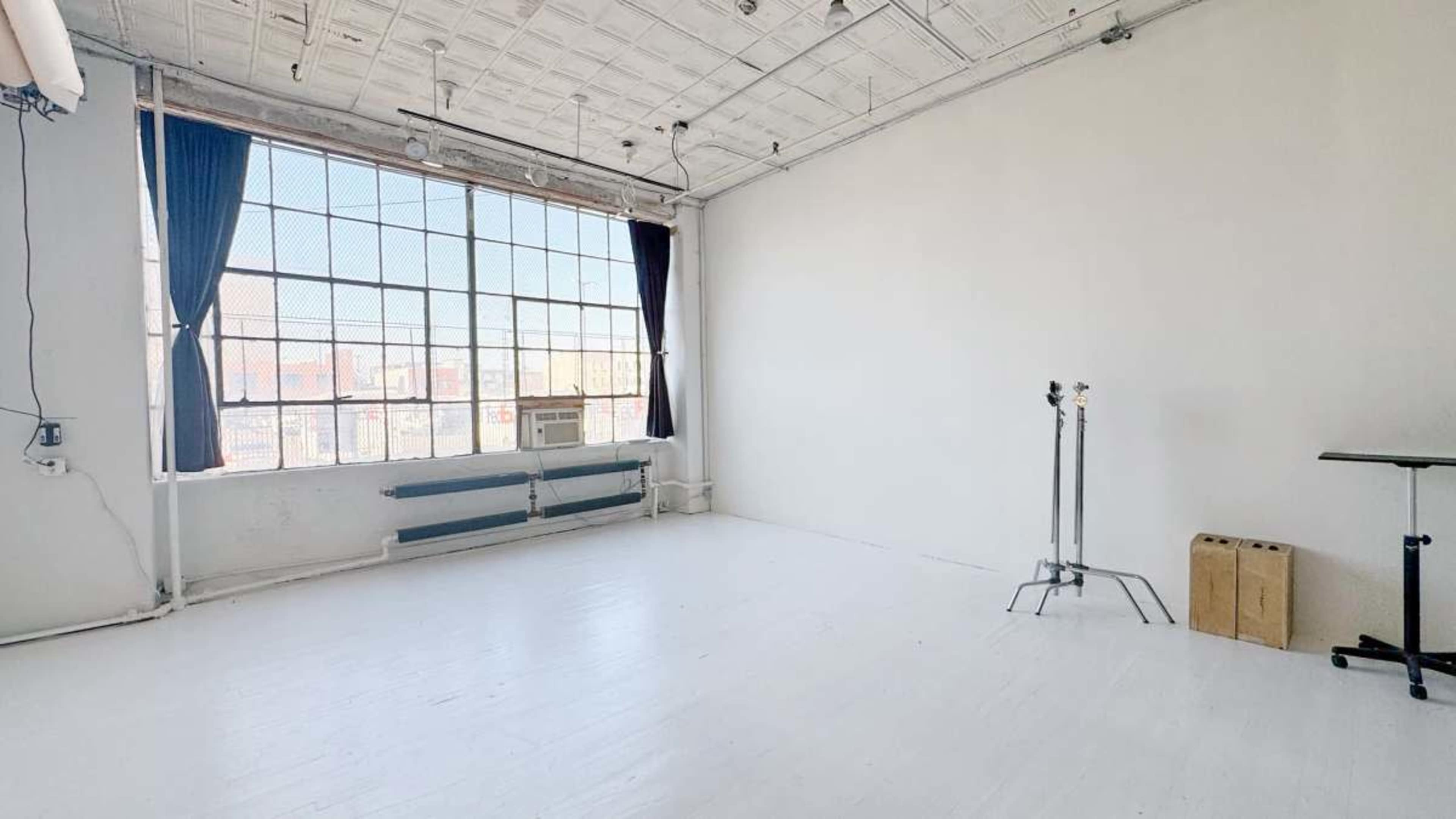 The image shows a spacious, empty room with large windows, white walls, and minimal furnishings including a standing table and a clothing rack.