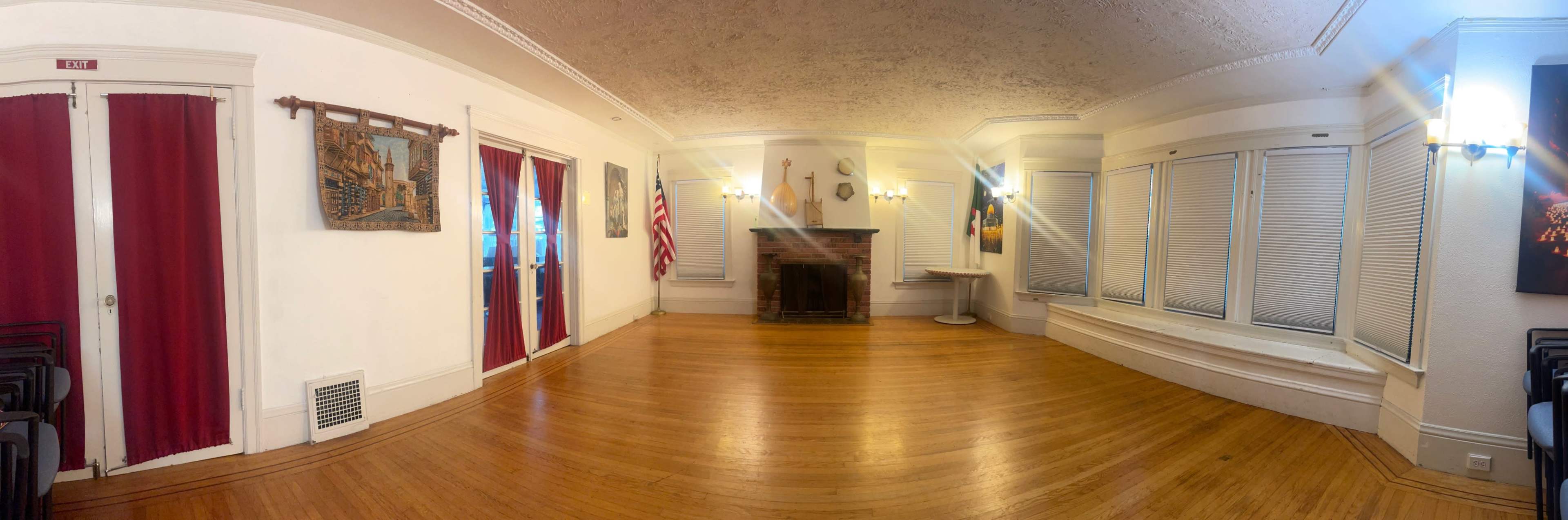 The image shows a spacious room with wooden flooring, a fireplace at the center, large windows, and a few wall decorations, including an American flag and artwork.