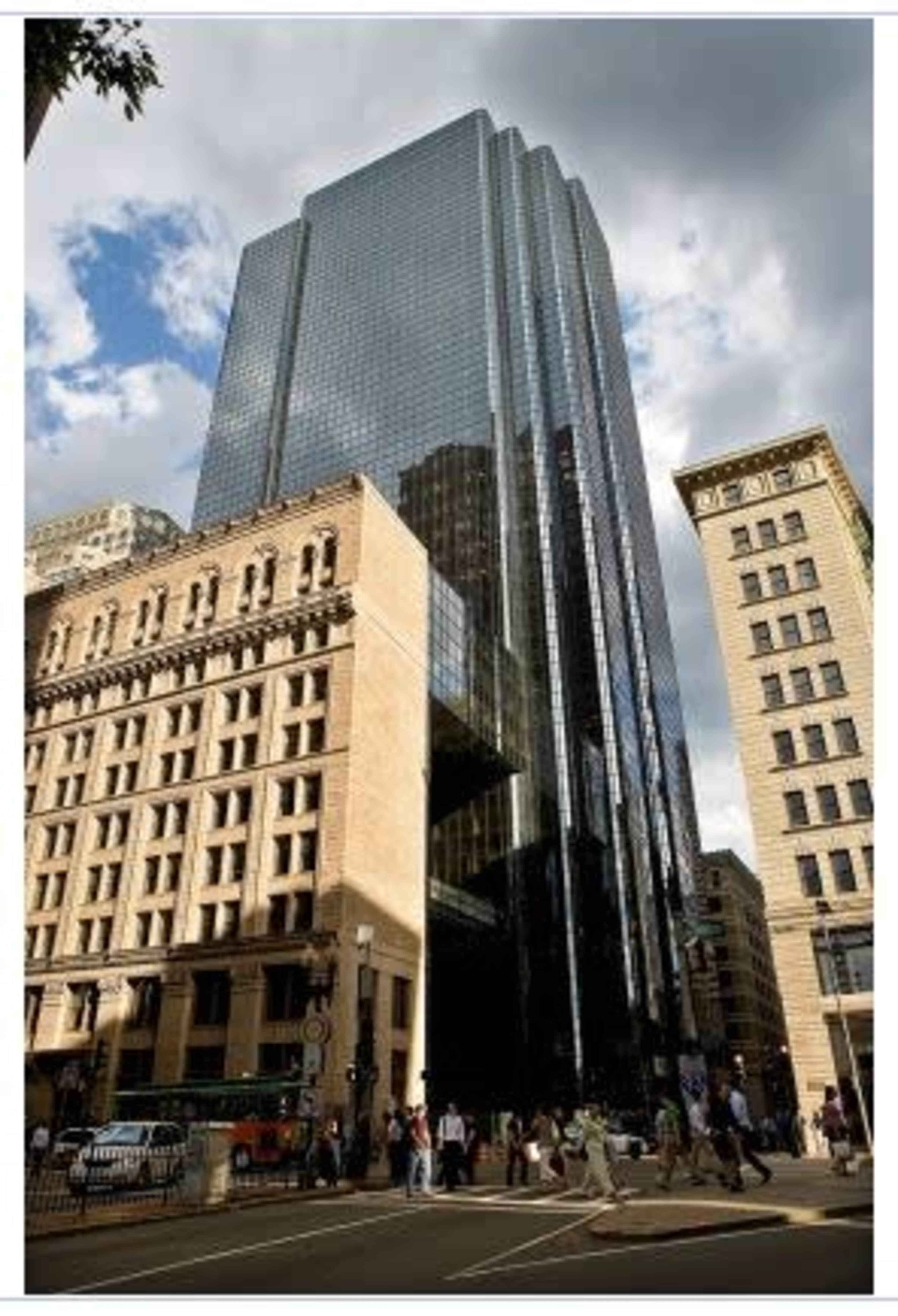 A modern glass skyscraper stands adjacent to a historic stone building, with pedestrians walking along the street below under a partly cloudy sky.