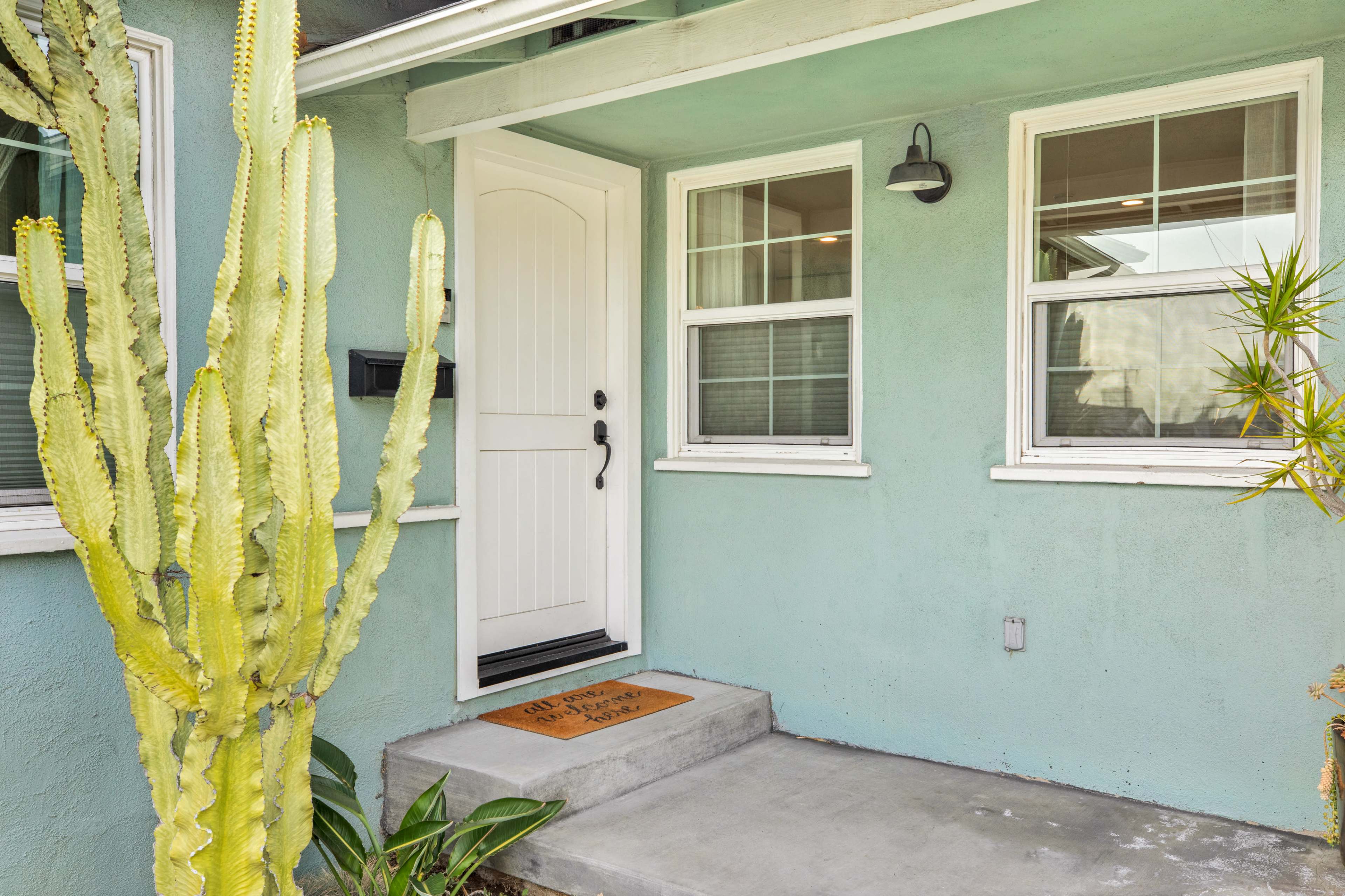 The image shows a light blue house with a white front door and several windows, accompanied by a tall cactus and a small welcome mat on the concrete steps.