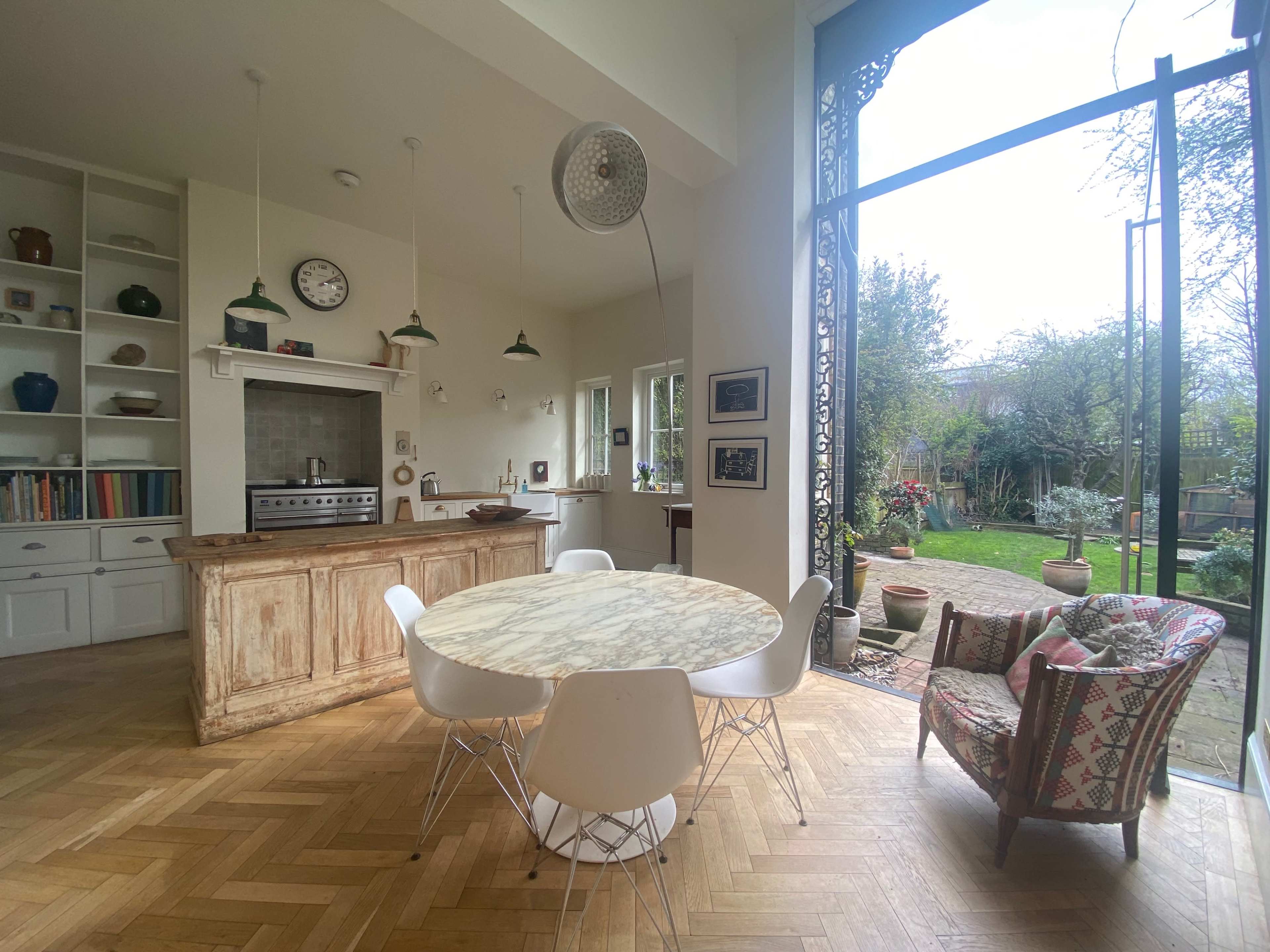 A spacious kitchen with a marble-top dining table, surrounded by white chairs, opens up to a garden through large glass doors.