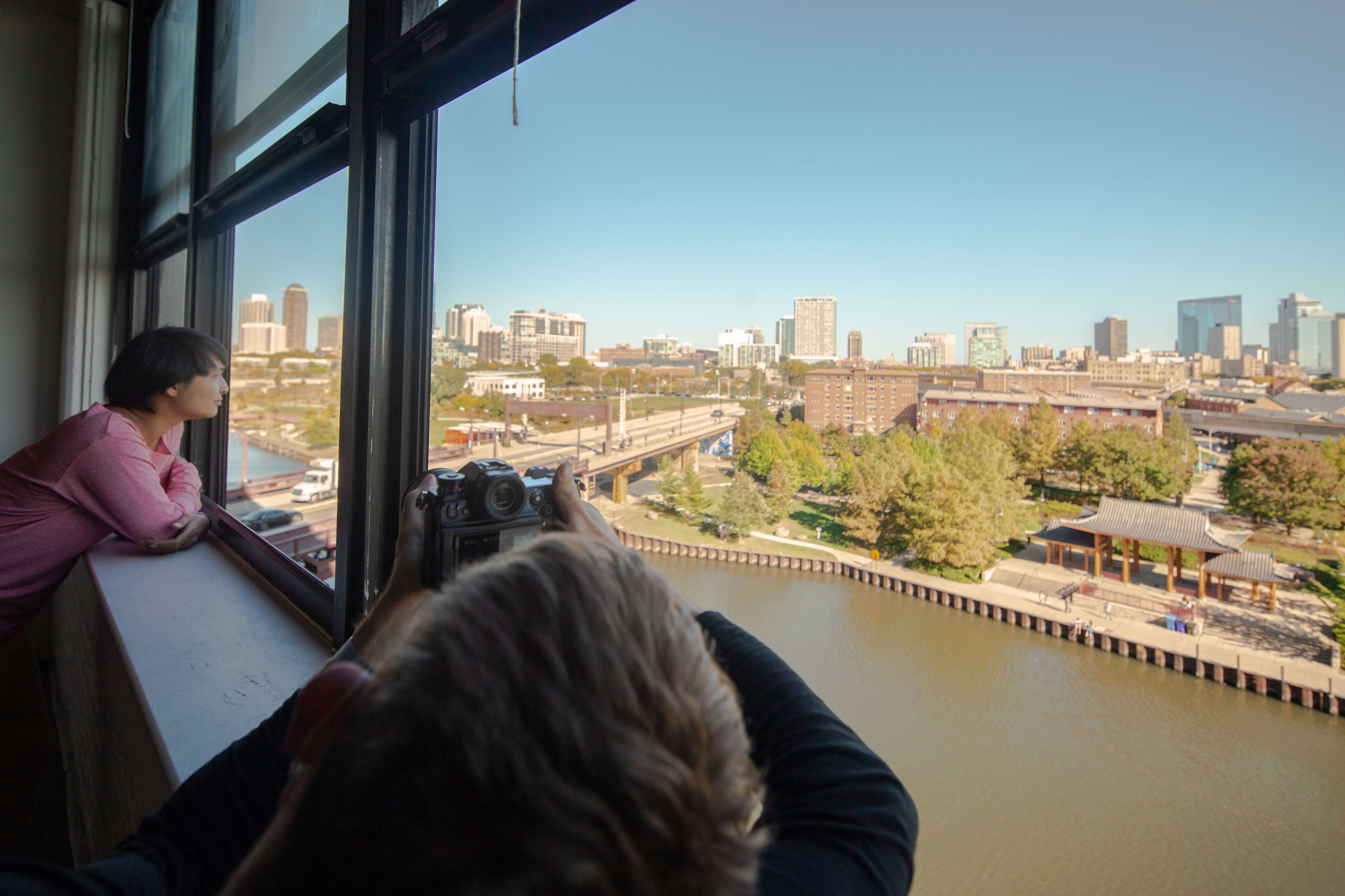 A person looks out the window while another takes a photo of the city skyline along a river.
