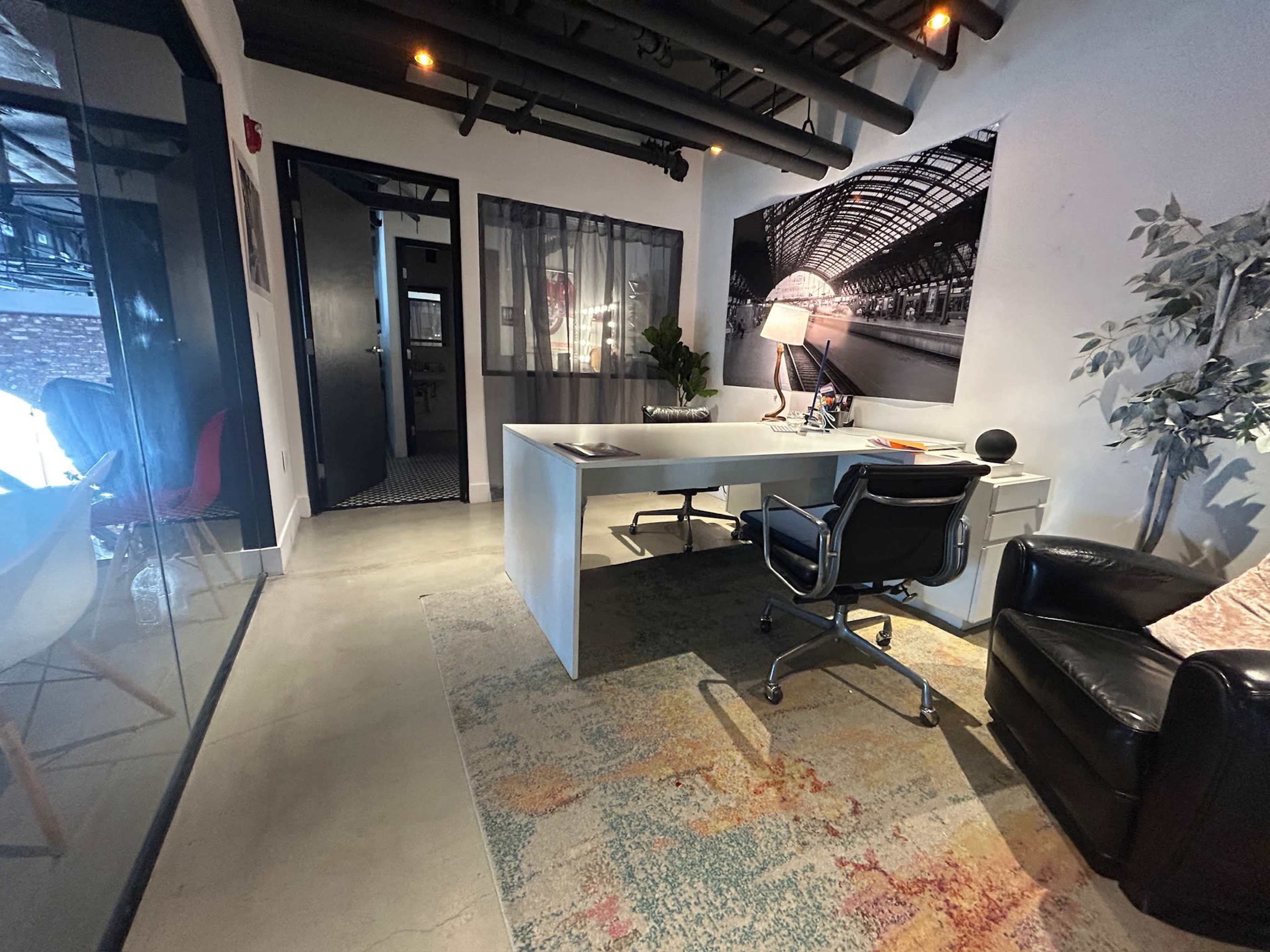 The image shows a modern office space featuring a desk with a chair, a black leather armchair, and decorative elements like a plant and artwork on the walls.