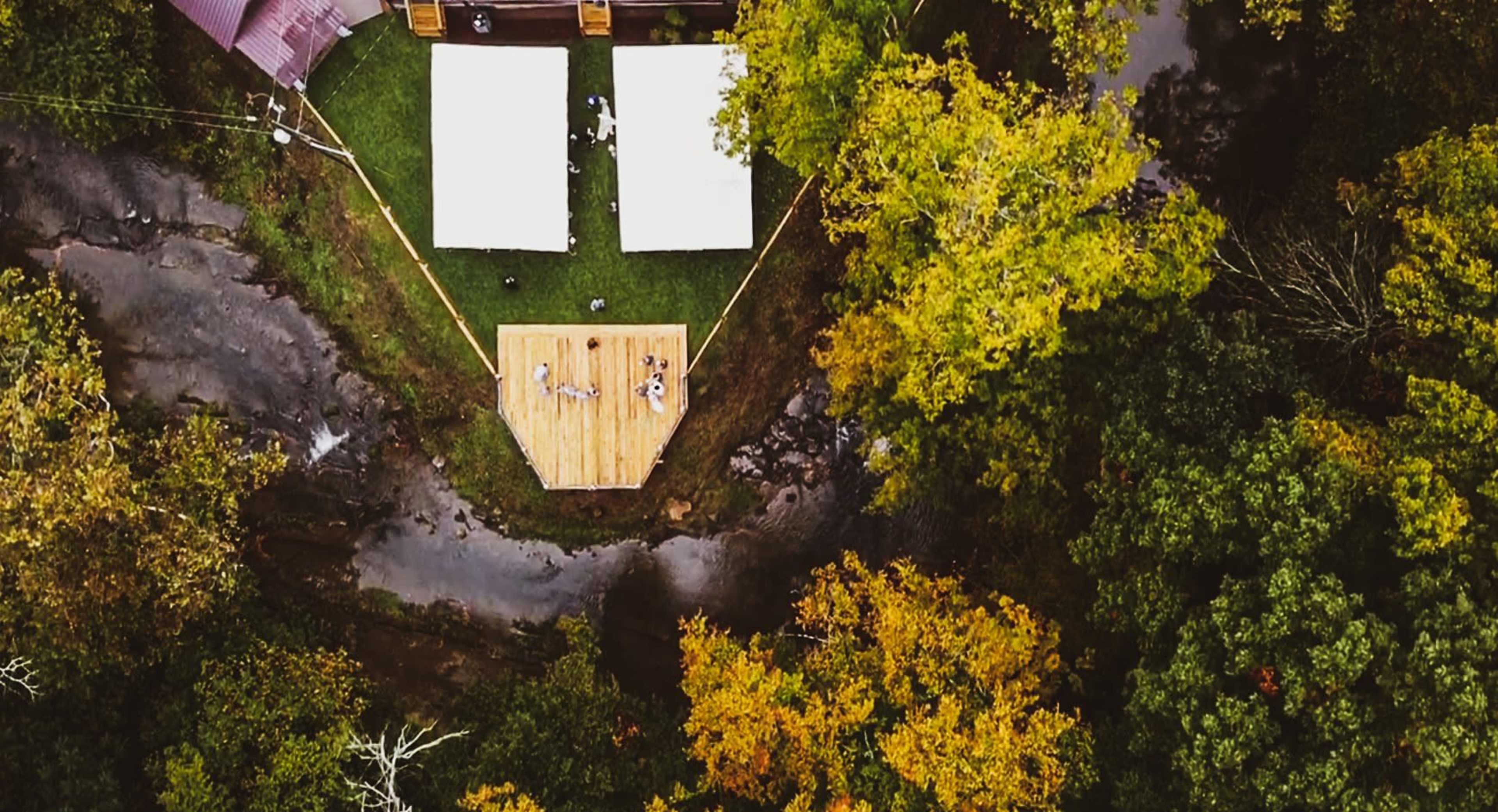 An aerial view shows a wooden deck surrounded by a creek and autumn-colored trees, with white tents set up on the lawn.