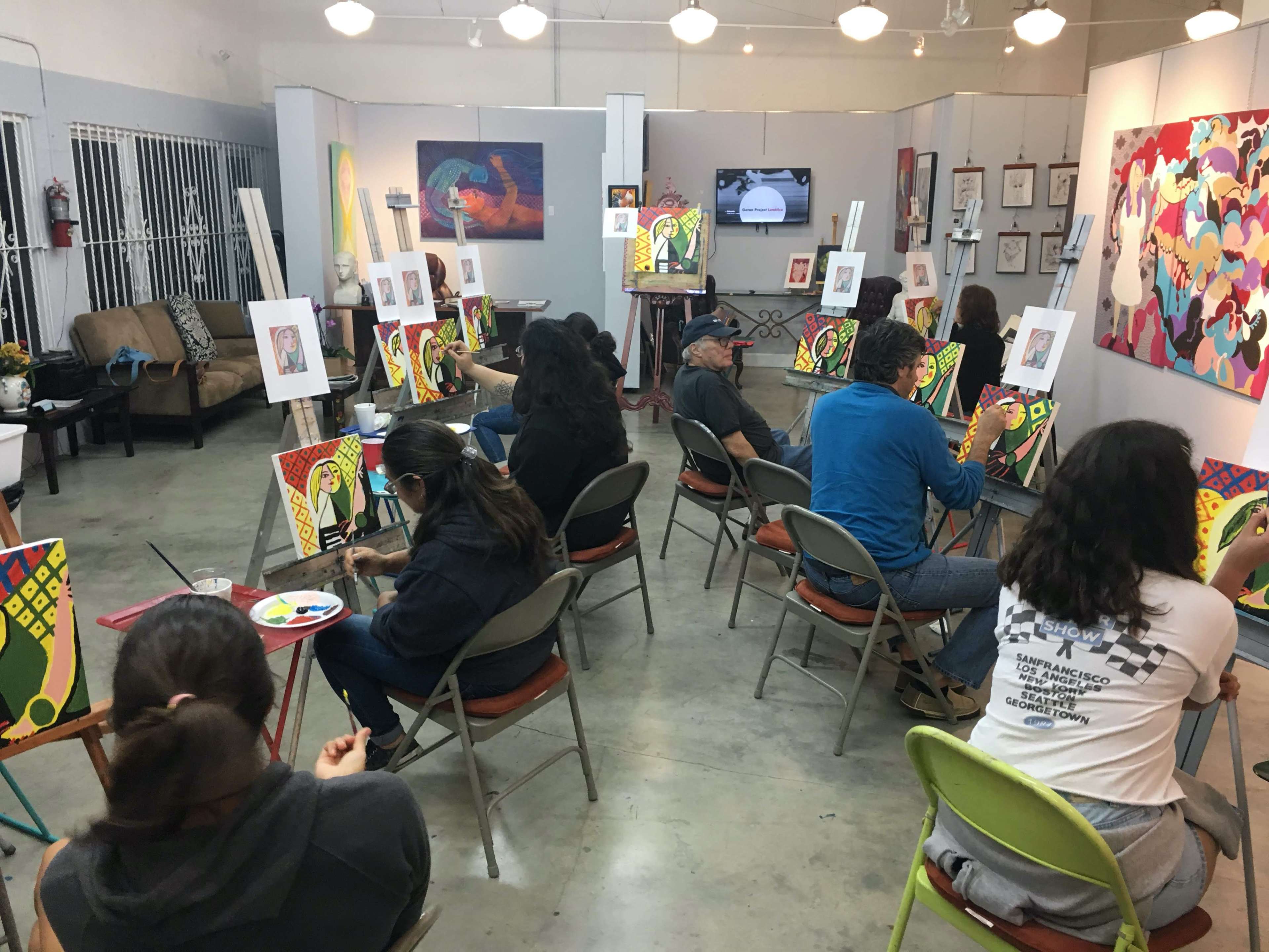 A group of people is engaged in a painting class within an art studio, with each person working on their own canvas while seated at folding chairs.