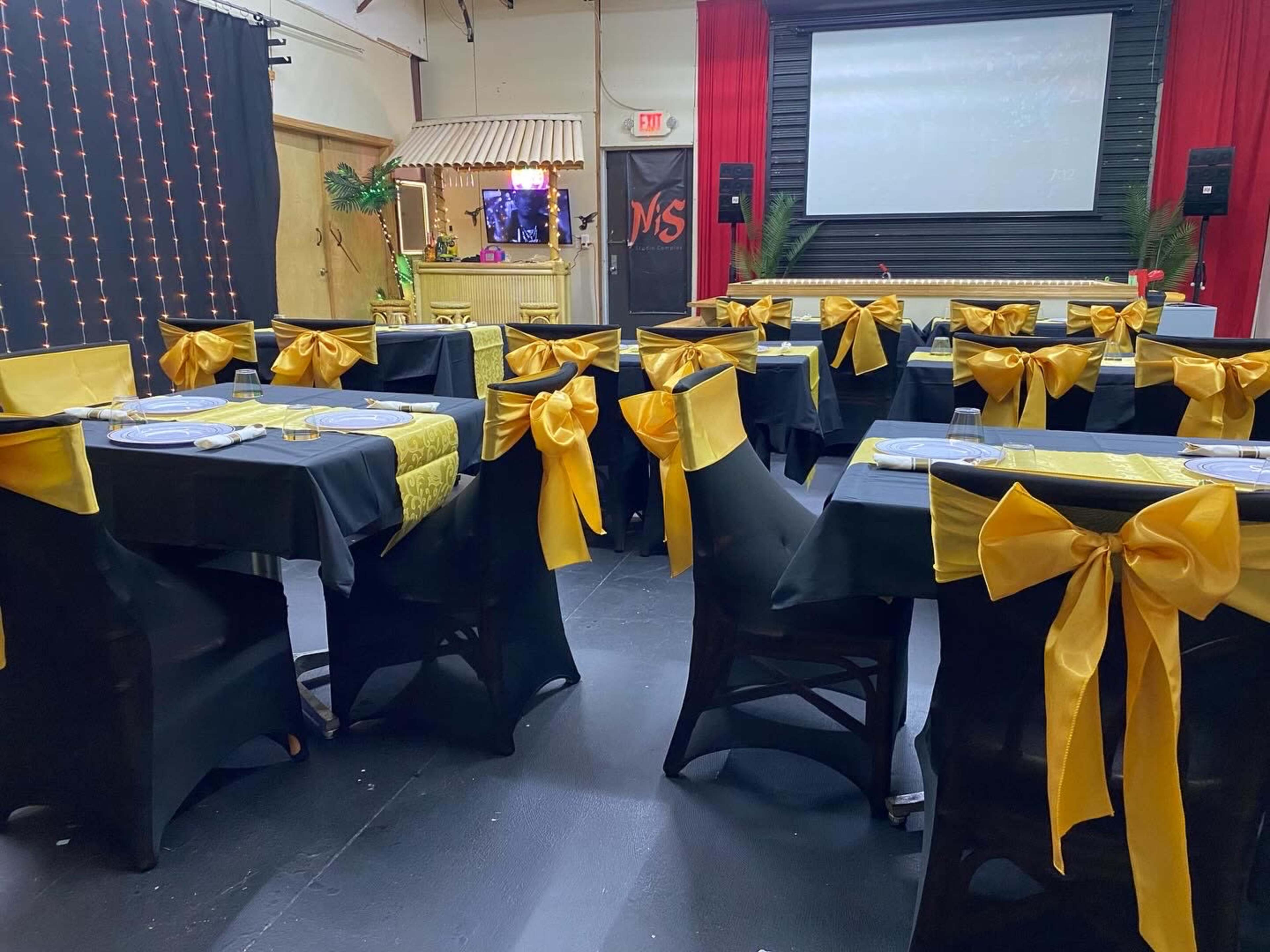 The image shows a banquet hall arranged with tables covered in black and gold tablecloths, each adorned with yellow bows on the chairs, alongside a projector screen displaying a presentation.