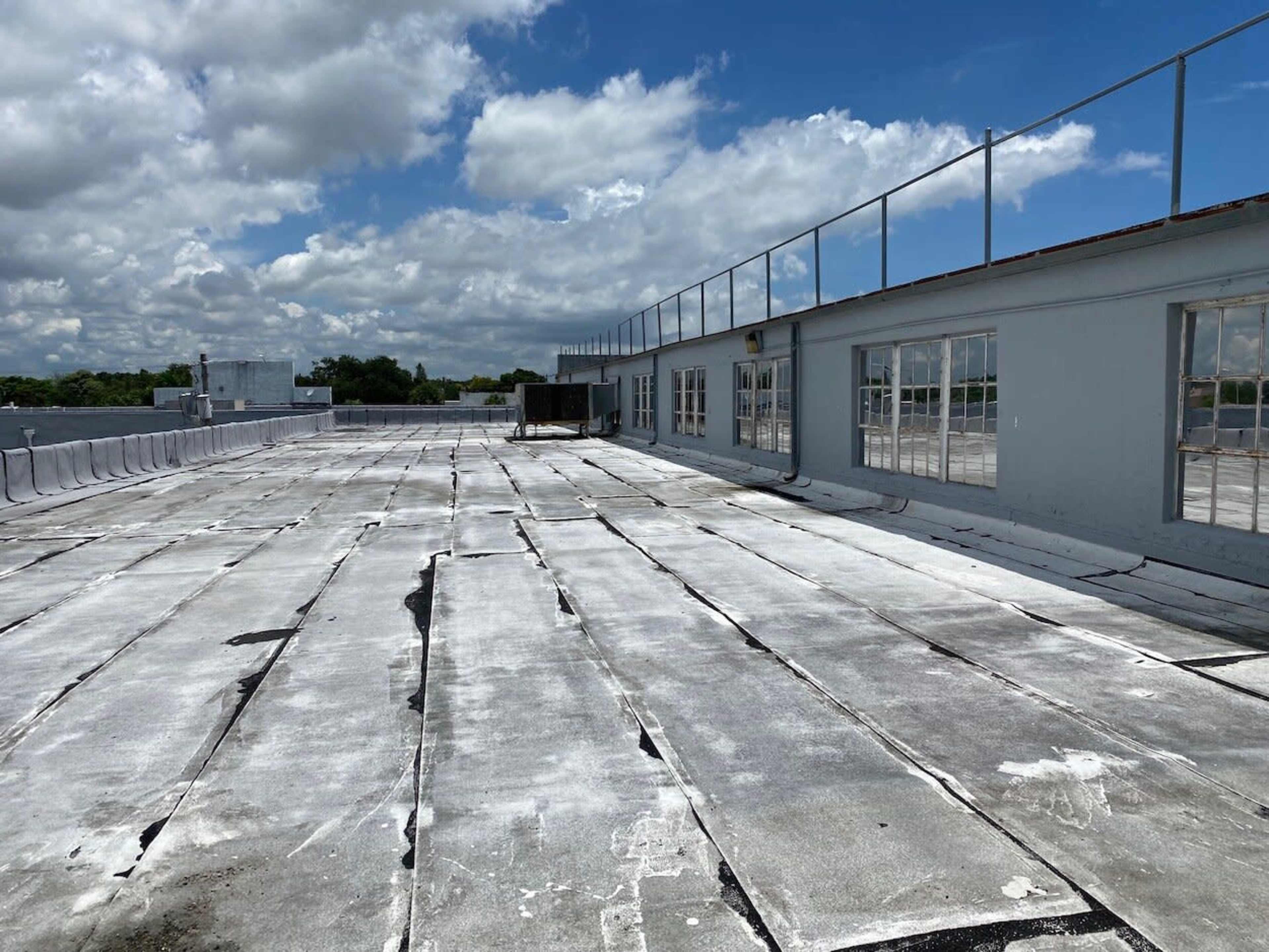 The image shows a flat rooftop with a cracked surface, surrounded by a low railing and a building with large windows under a partly cloudy sky.