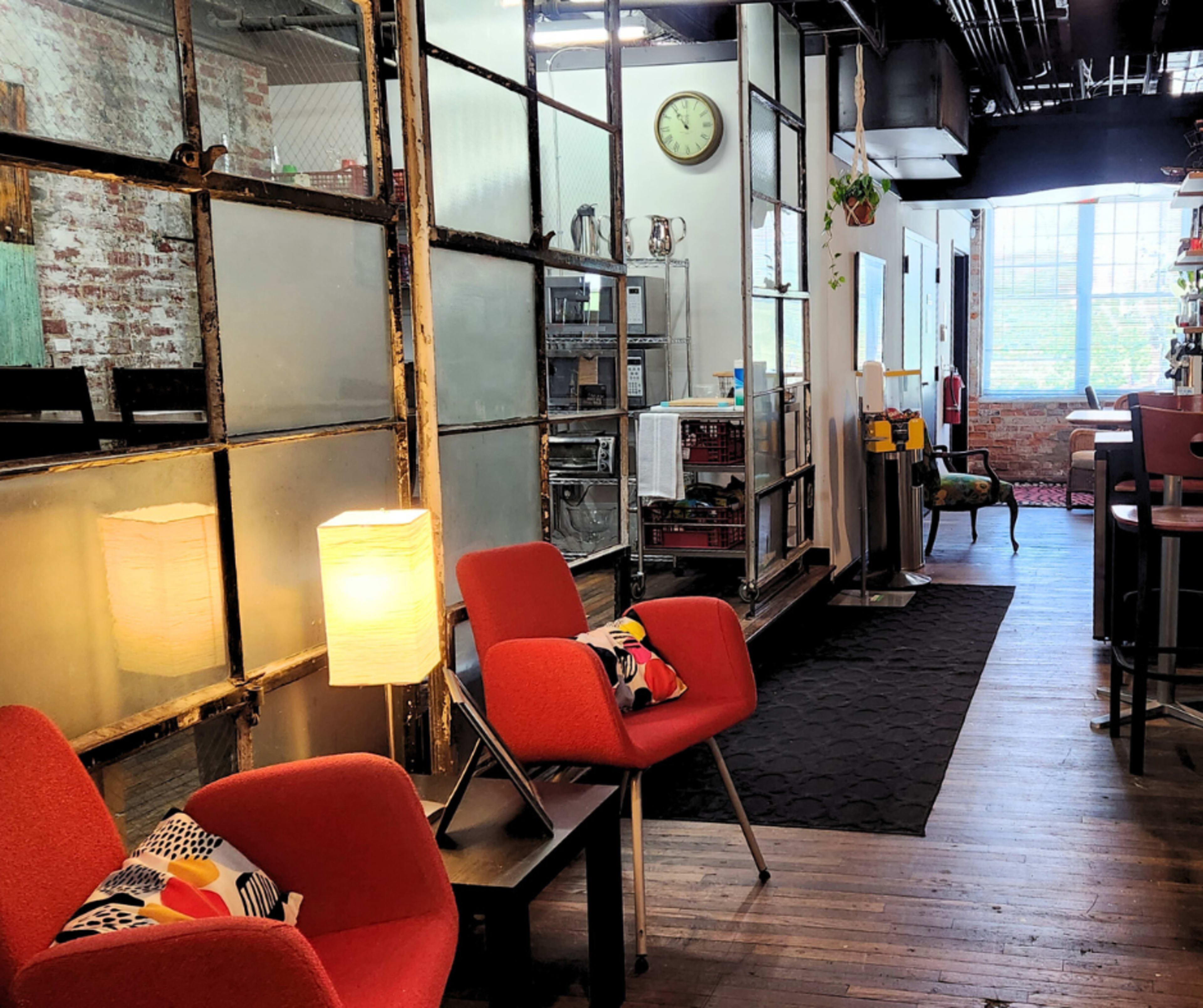 The image shows a modern workspace featuring red chairs, a lamp, and large windows with industrial-looking glass partitions.