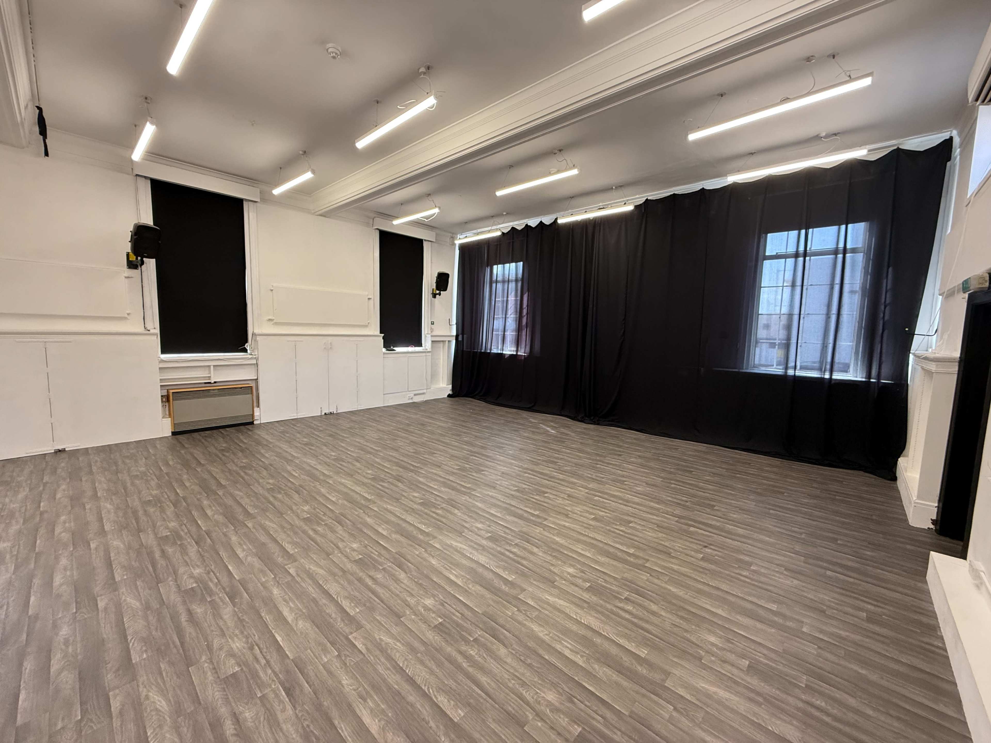 The image shows an empty room with a wooden floor, large windows covered by black curtains, and minimal furnishings.