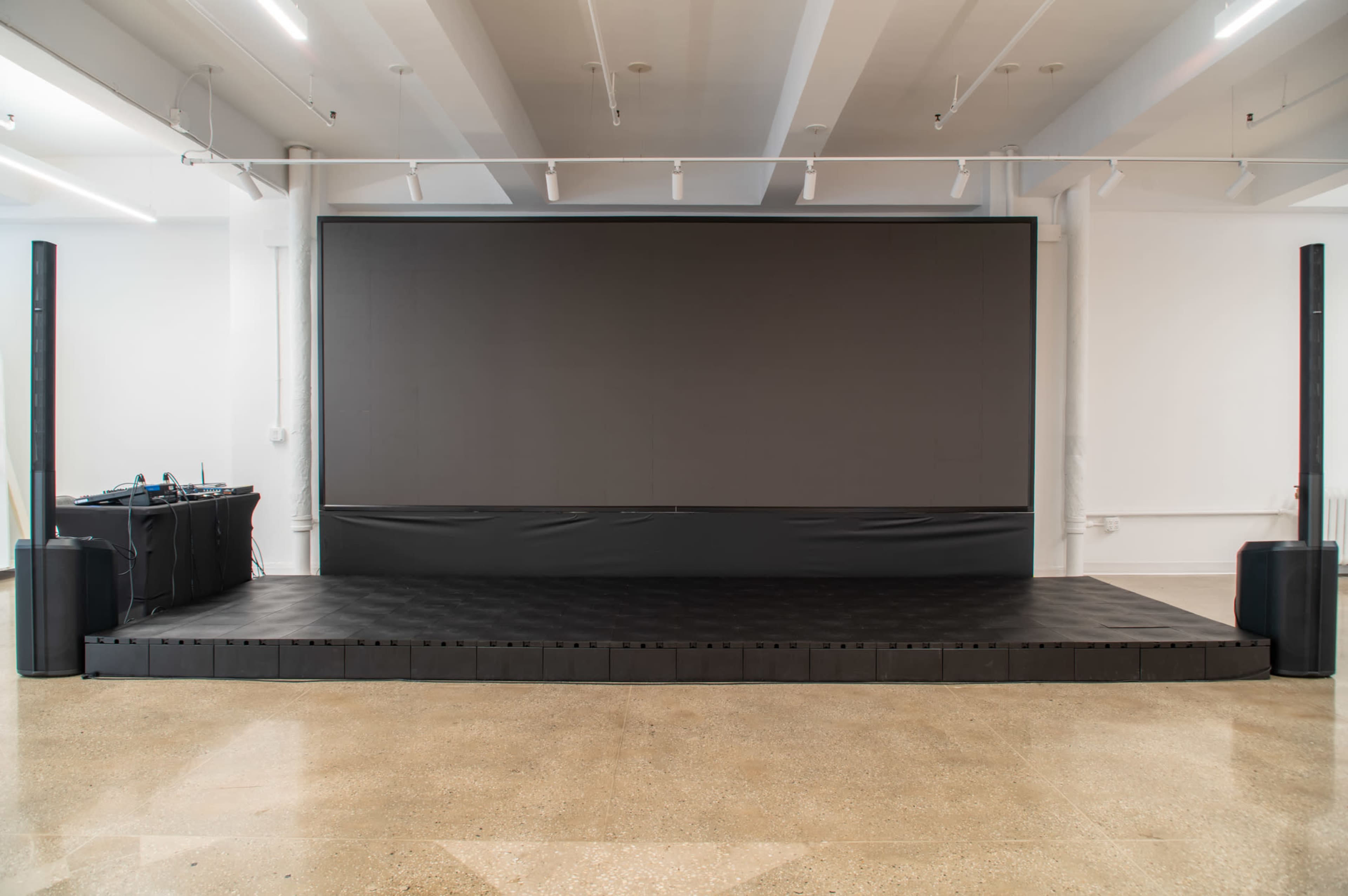 The image shows a spacious indoor area featuring a large blank screen, a raised black stage, and two vertical speaker columns on either side.