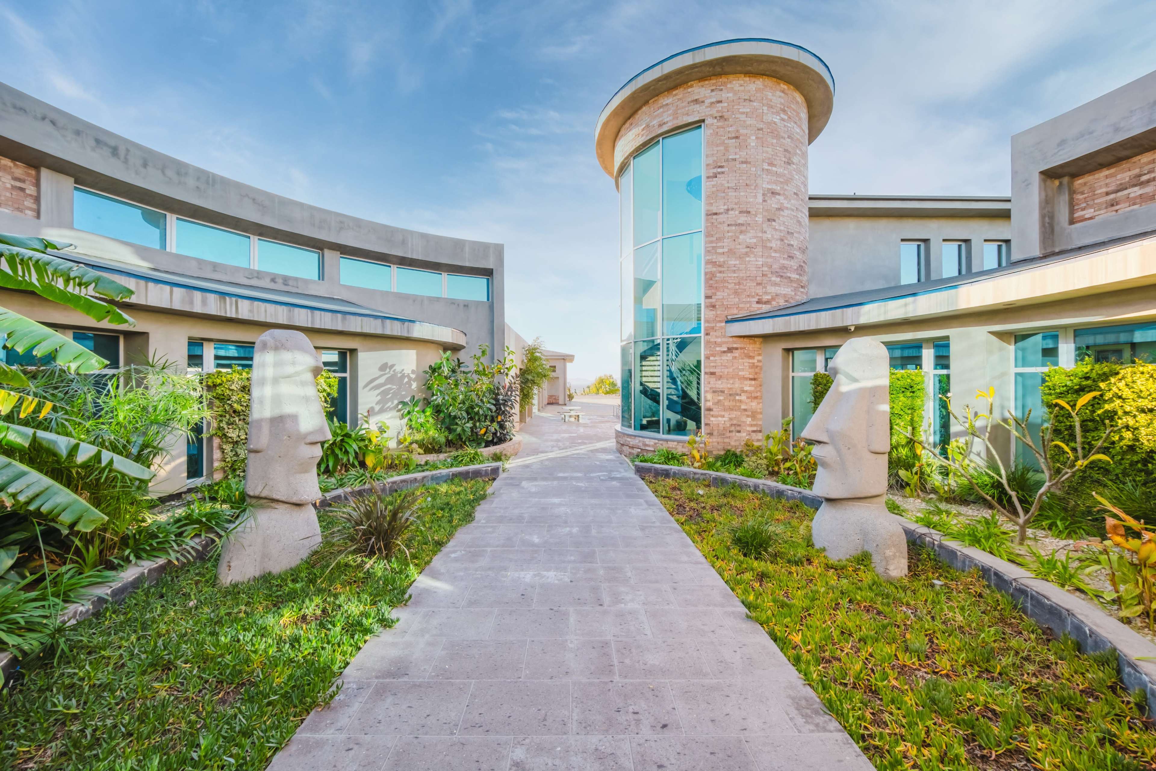 The image shows a modern architectural complex with a central pathway flanked by two large stone sculptures resembling Easter Island heads, surrounded by greenery and a clear blue sky.