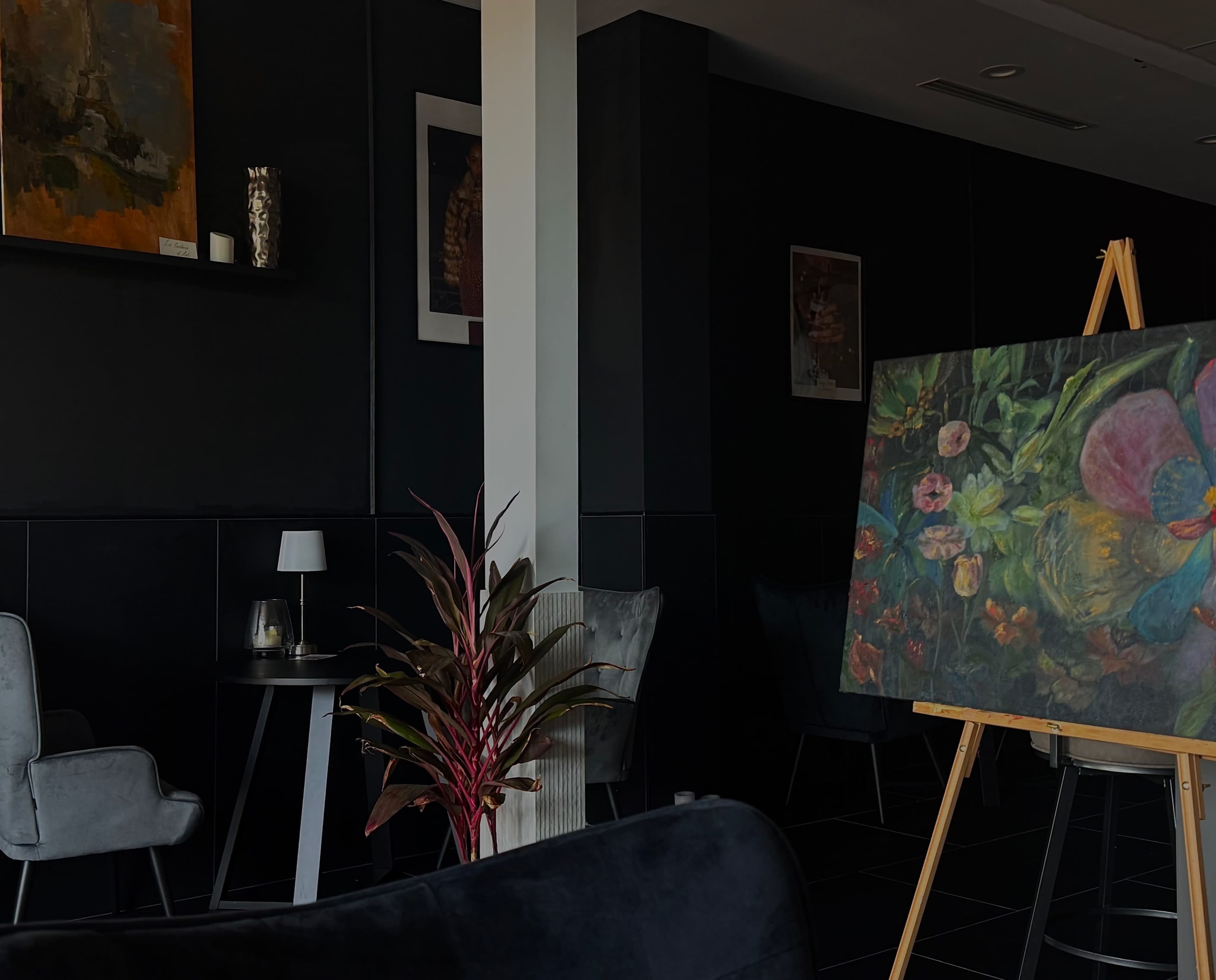 The image shows a darkly lit café with a large floral painting on an easel, surrounded by stylish seating and decorative elements.