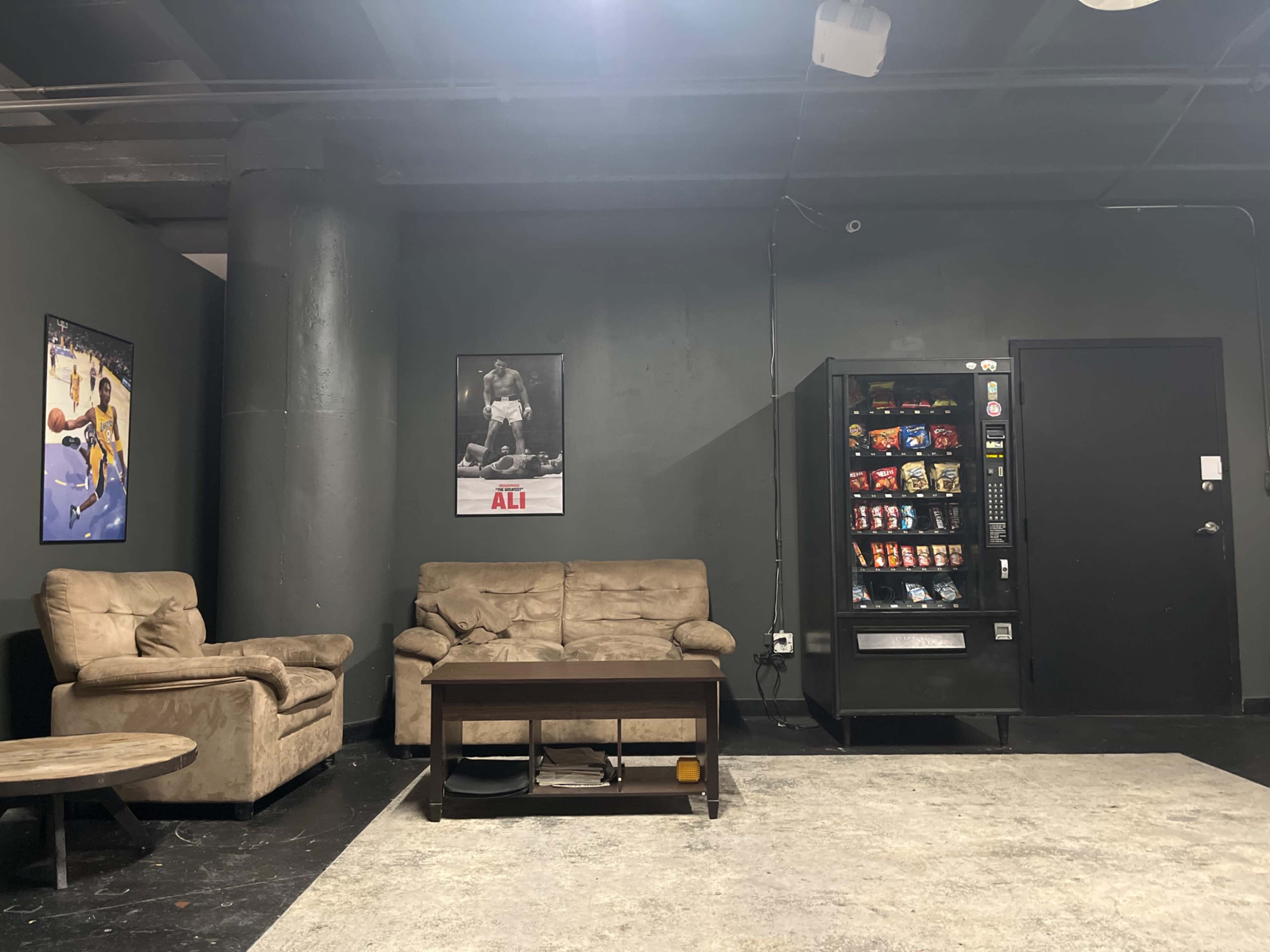 A room with two beige armchairs, a coffee table, a vending machine filled with snacks and drinks, and posters on the walls, including one featuring a famous boxer.