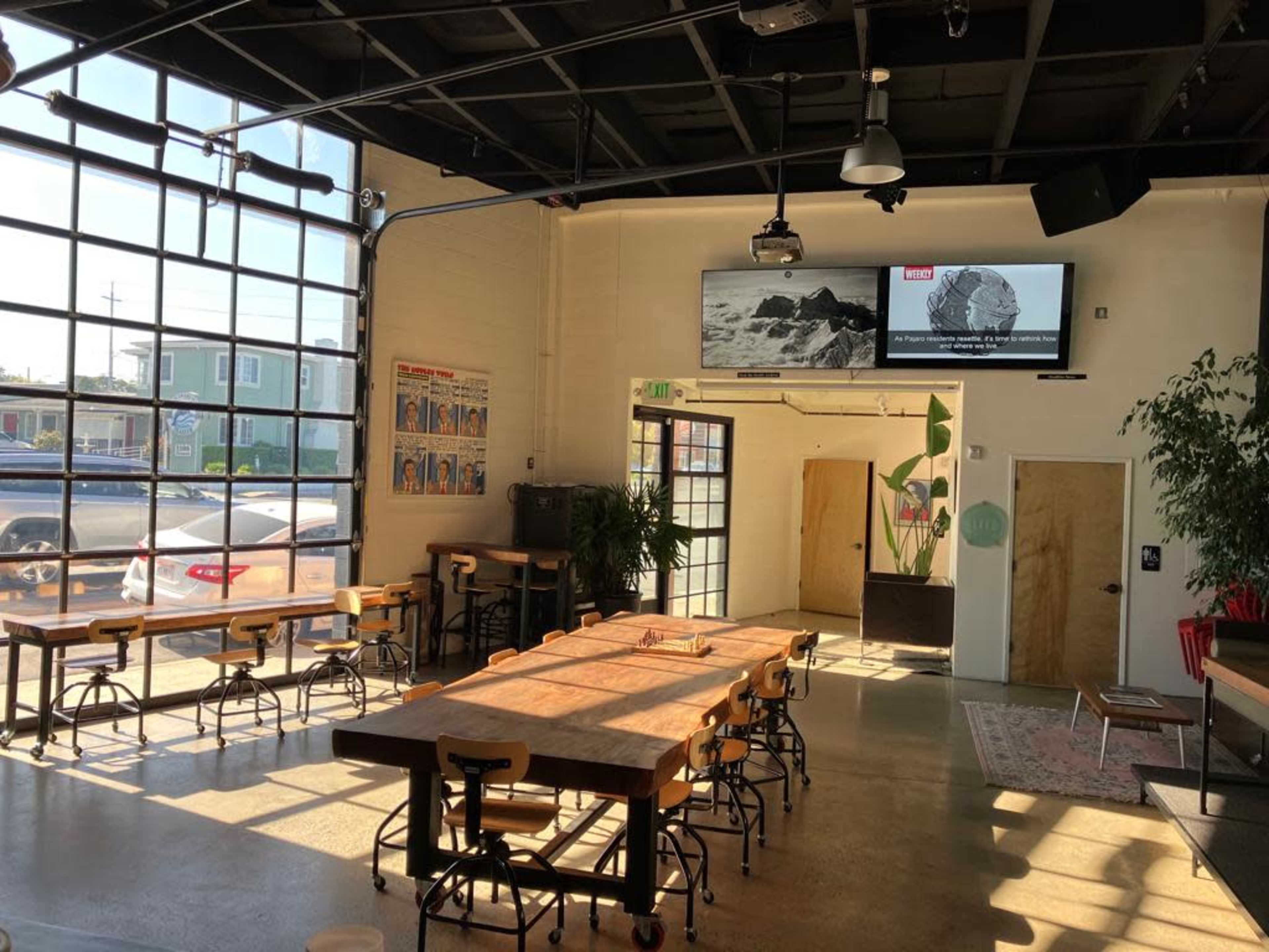 The interior of a spacious café features large windows, several wooden tables, and a screen displaying an image on the wall.