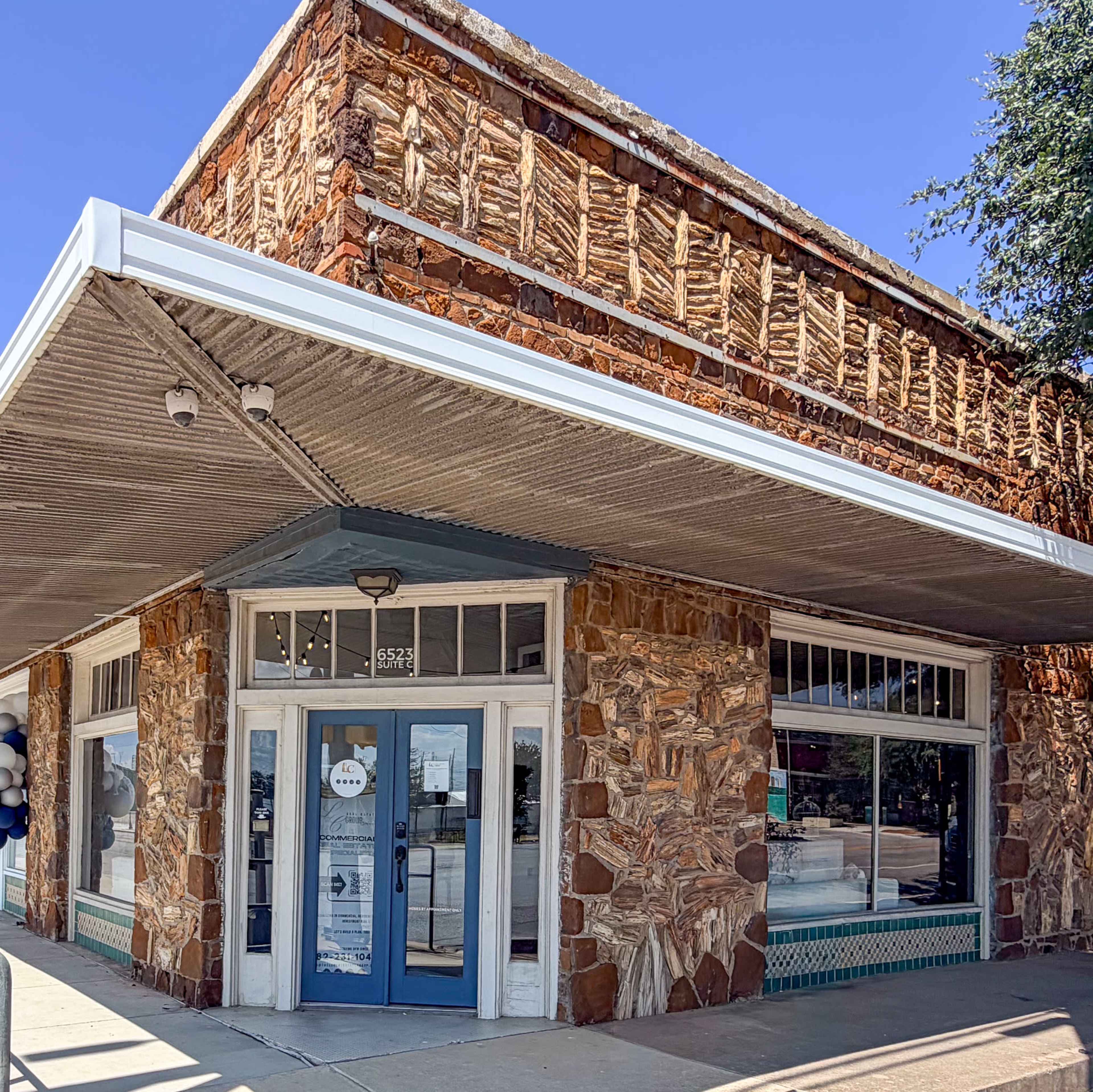 The image shows a building with a stone facade and a blue door, featuring large front windows and an overhanging roof.