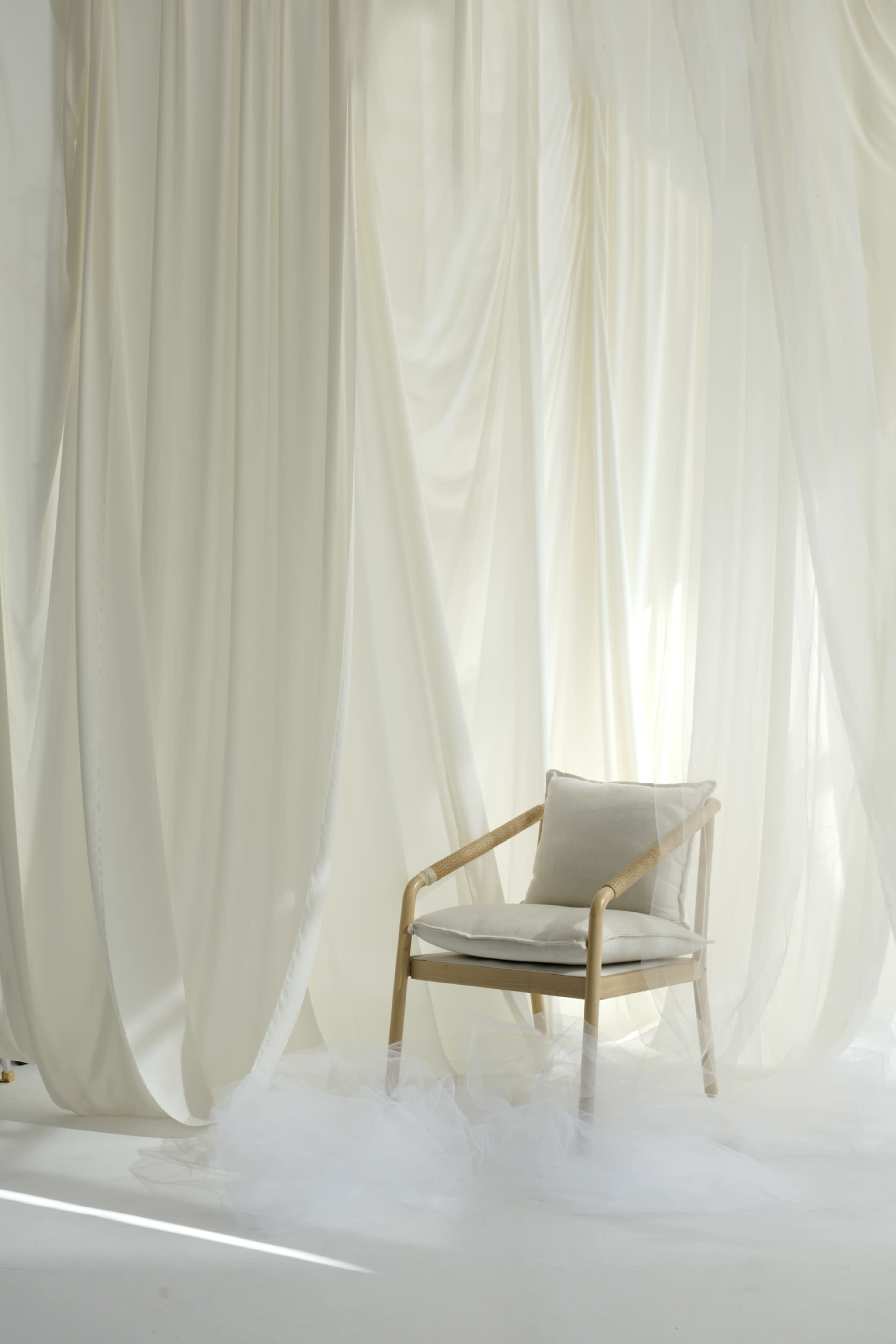 A wooden chair with a cushion is placed in front of sheer white fabric drapes, with a misty white covering the floor.