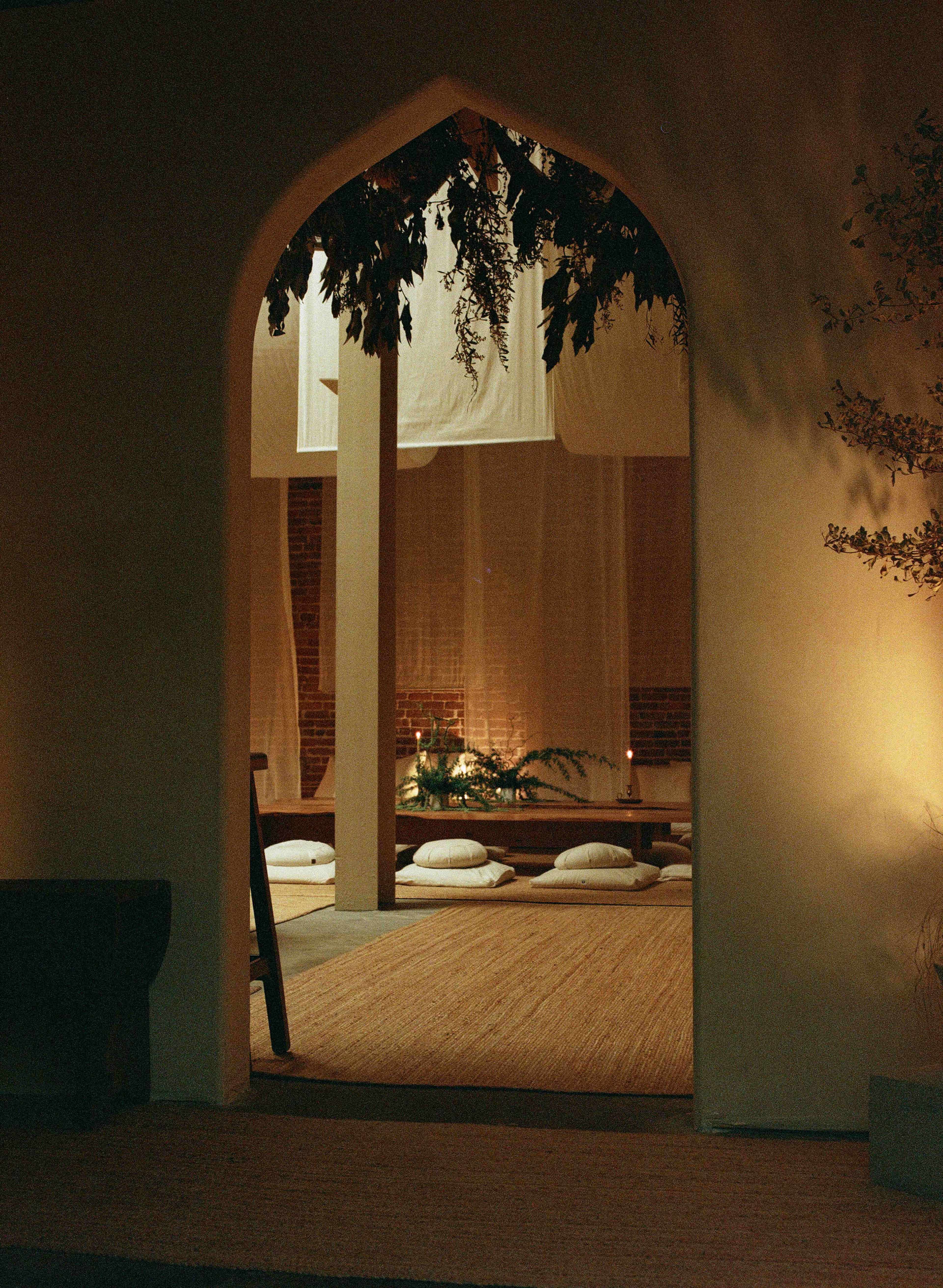 A serene indoor space features low seating cushions arranged around a central area, softly lit by candles and natural elements.