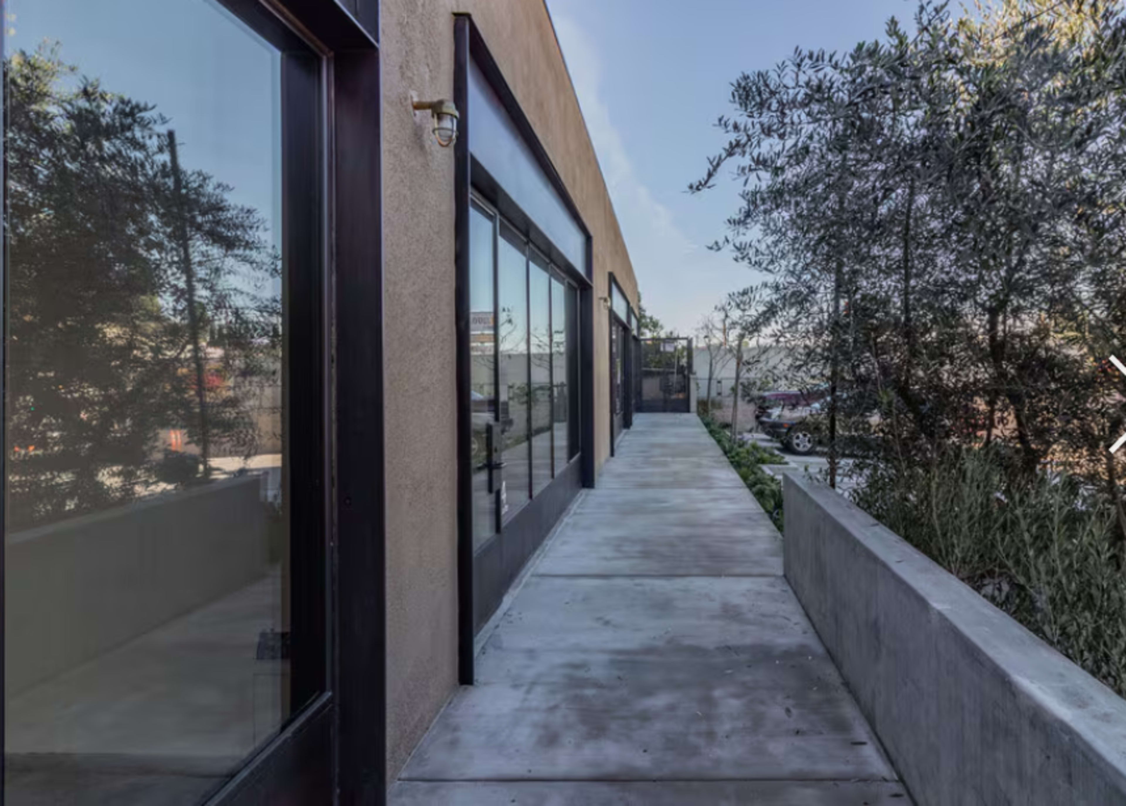 The image shows a concrete walkway alongside a building with large glass windows and greenery on one side.