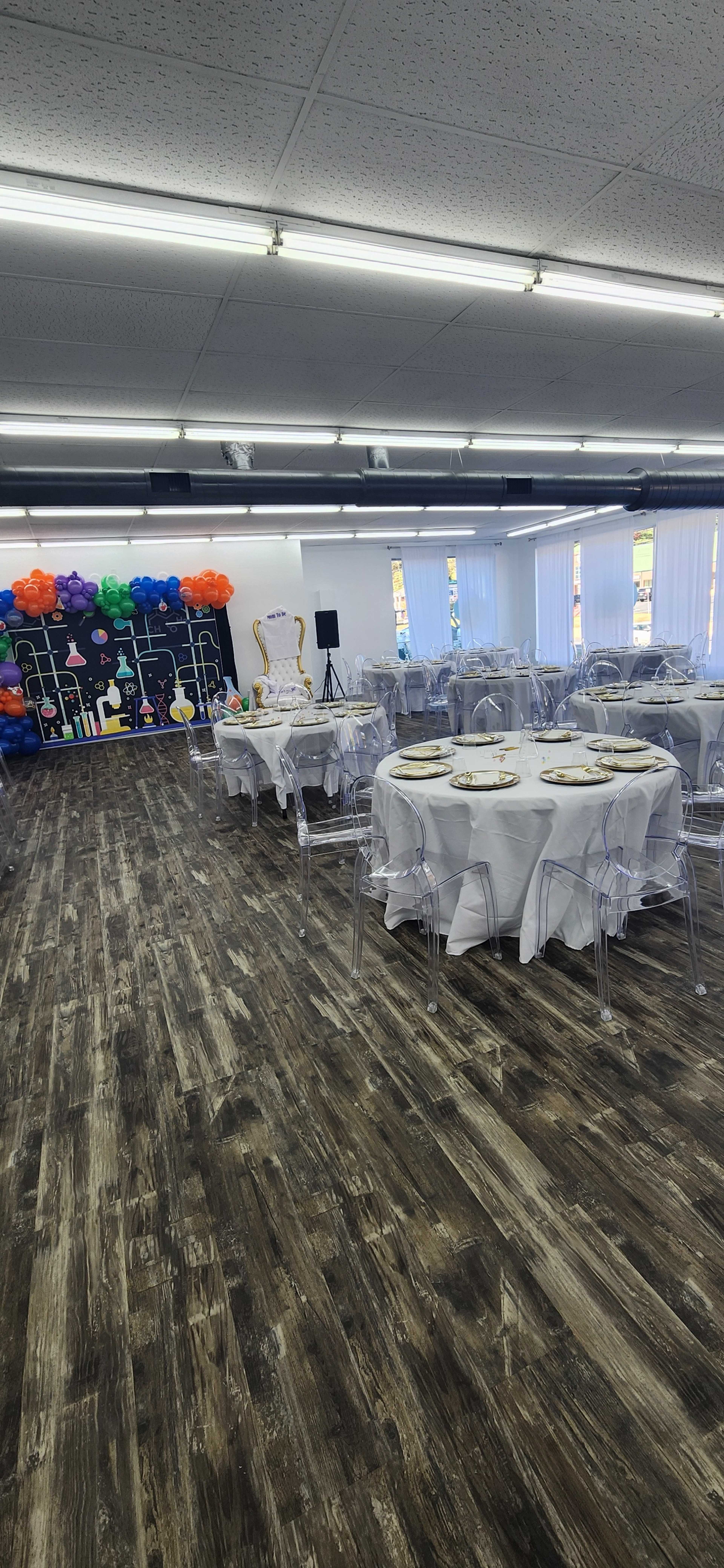 The image shows a banquet hall set up for an event, featuring round tables with white tablecloths and transparent chairs, alongside a colorful backdrop of balloons.