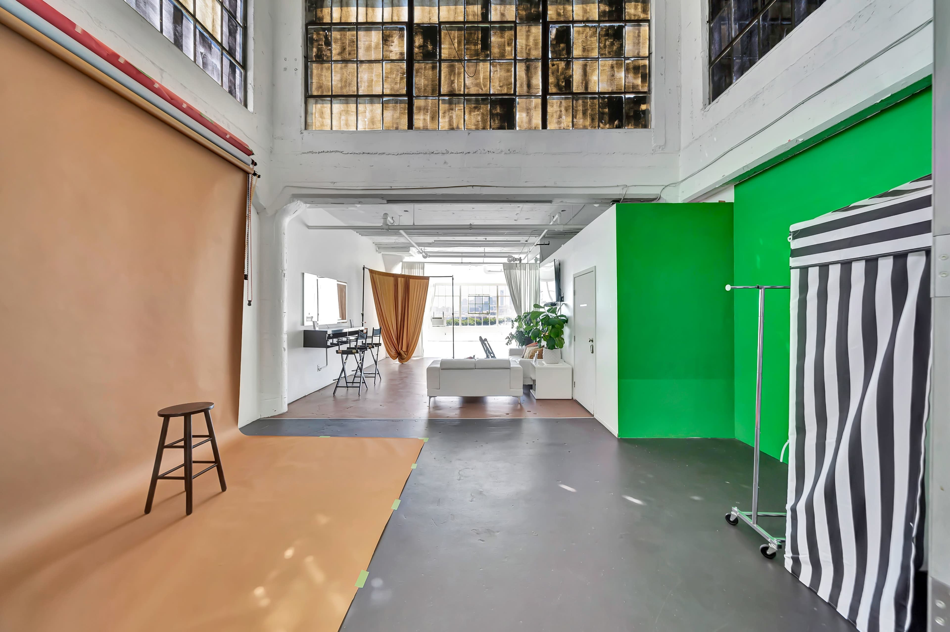 A spacious studio with a tan backdrop on one side, a green backdrop on another, and a striped partition, featuring large windows allowing natural light to enter.