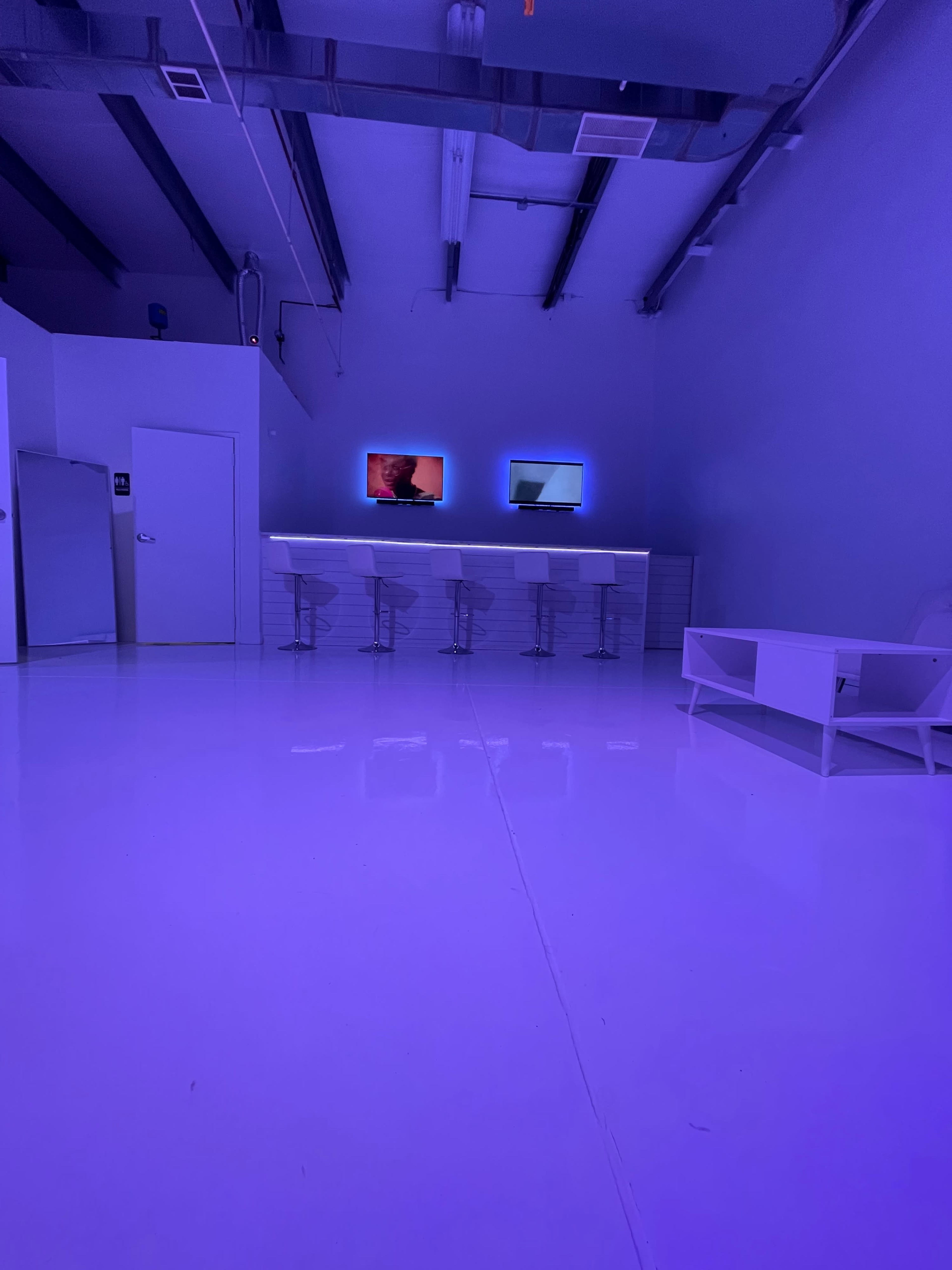 A brightly lit, purple-toned studio features a bar with stools and two screens on the wall, set against a glossy white floor.