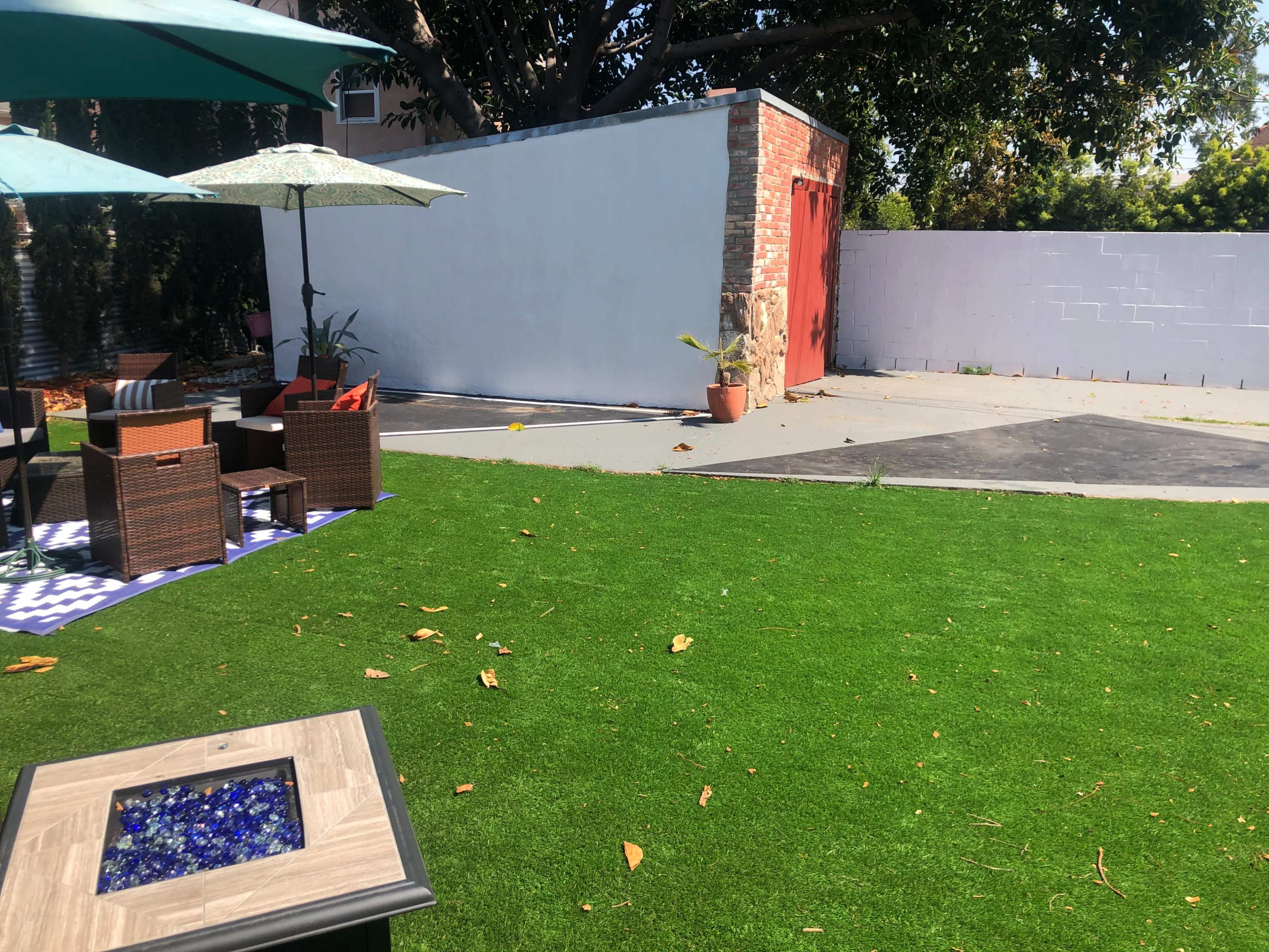 The scene features a grassy backyard with outdoor furniture, a storage shed with a red door, and a patio area shaded by umbrellas.